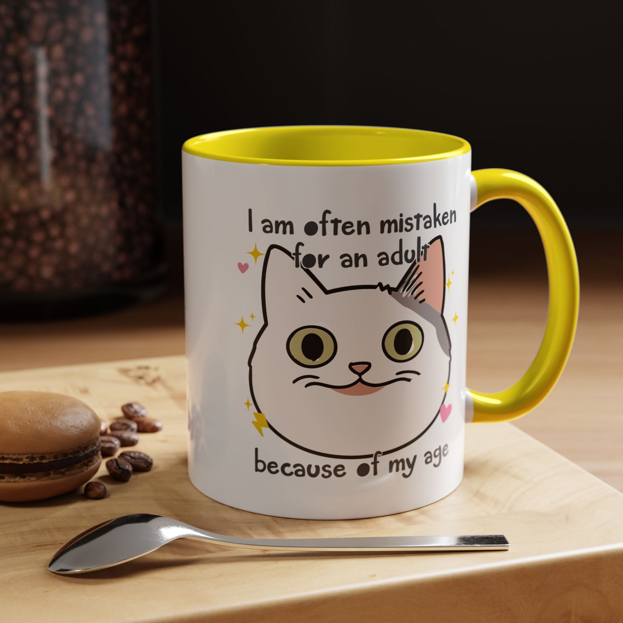 White ceramic mug with yellow handle and interior featuring a cute wide-eyed cat face, hearts, sparkles, and the phrase “I am often mistaken for an adult because of my age.”
