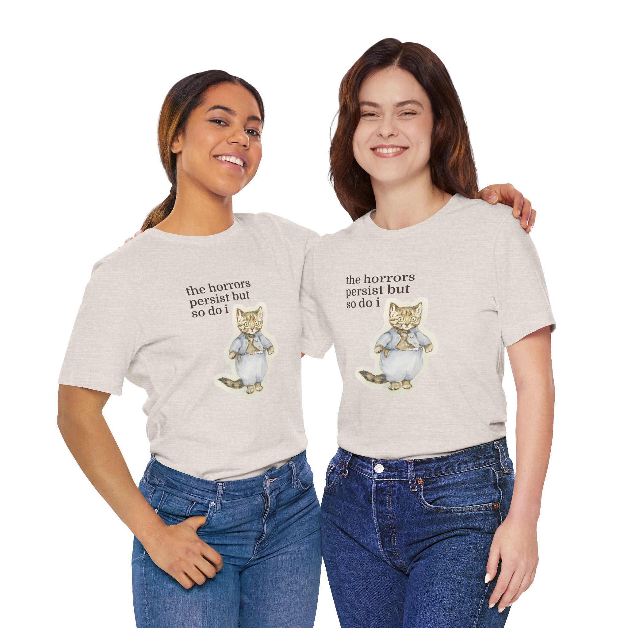 Two women wearing matching beige t-shirt featuring a vintage-style illustrated cat in blue clothing with the phrase “the horrors persist but so do i.”