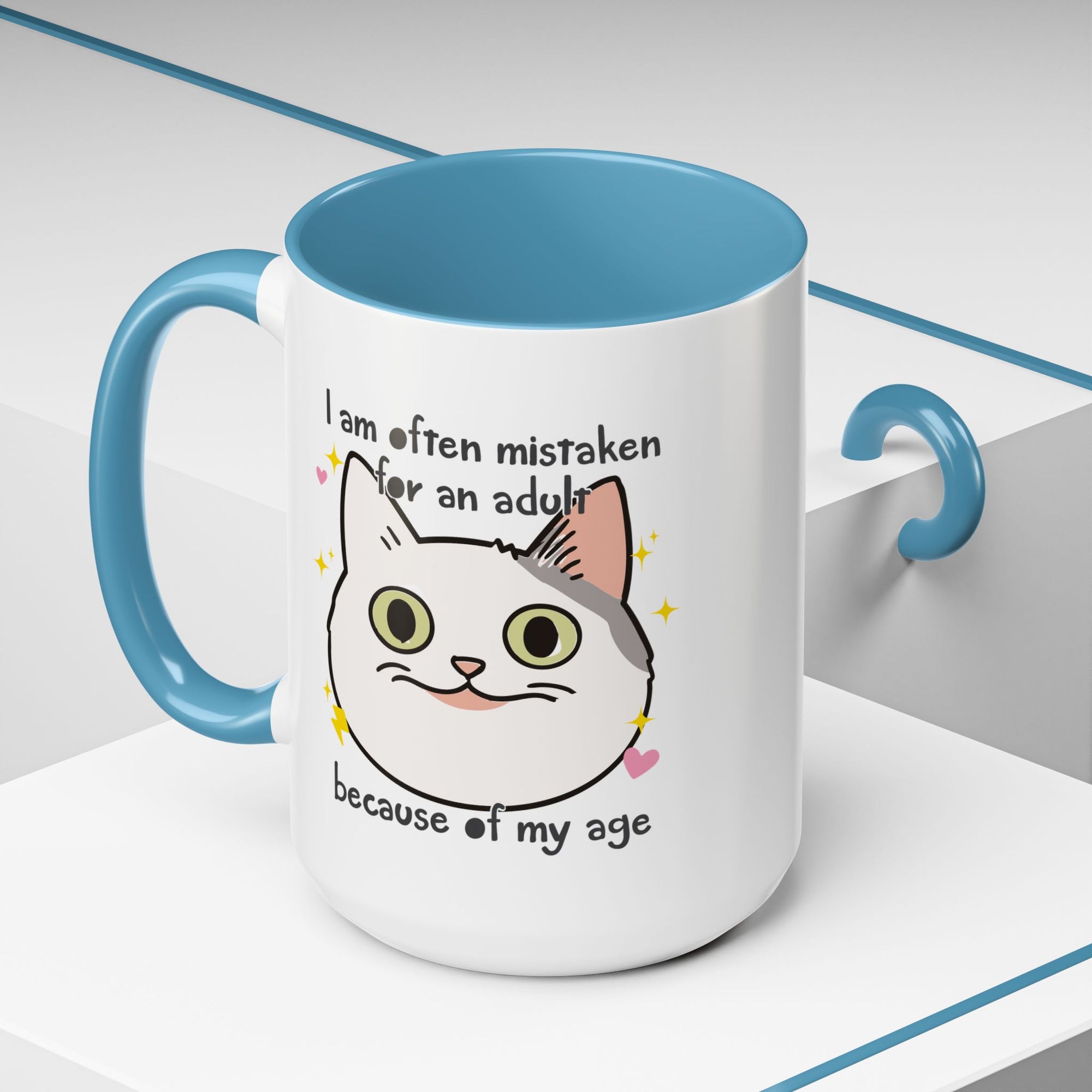 White ceramic mug with blue handle and interior featuring a cute wide-eyed cat face, hearts, sparkles, and the phrase “I am often mistaken for an adult because of my age.”

