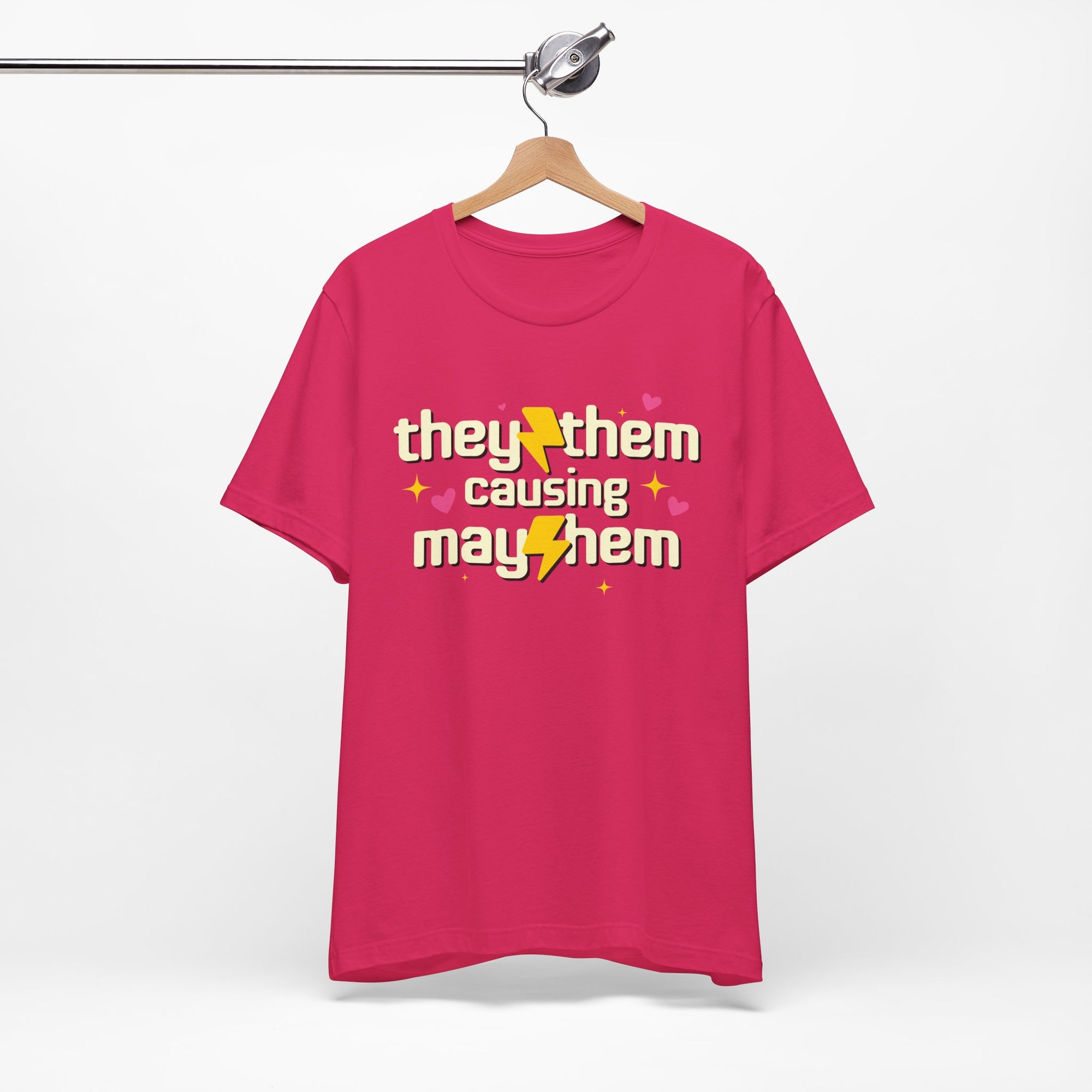 Pink t-shirt with 'they them causing mayhem' text on a hanger against a white background