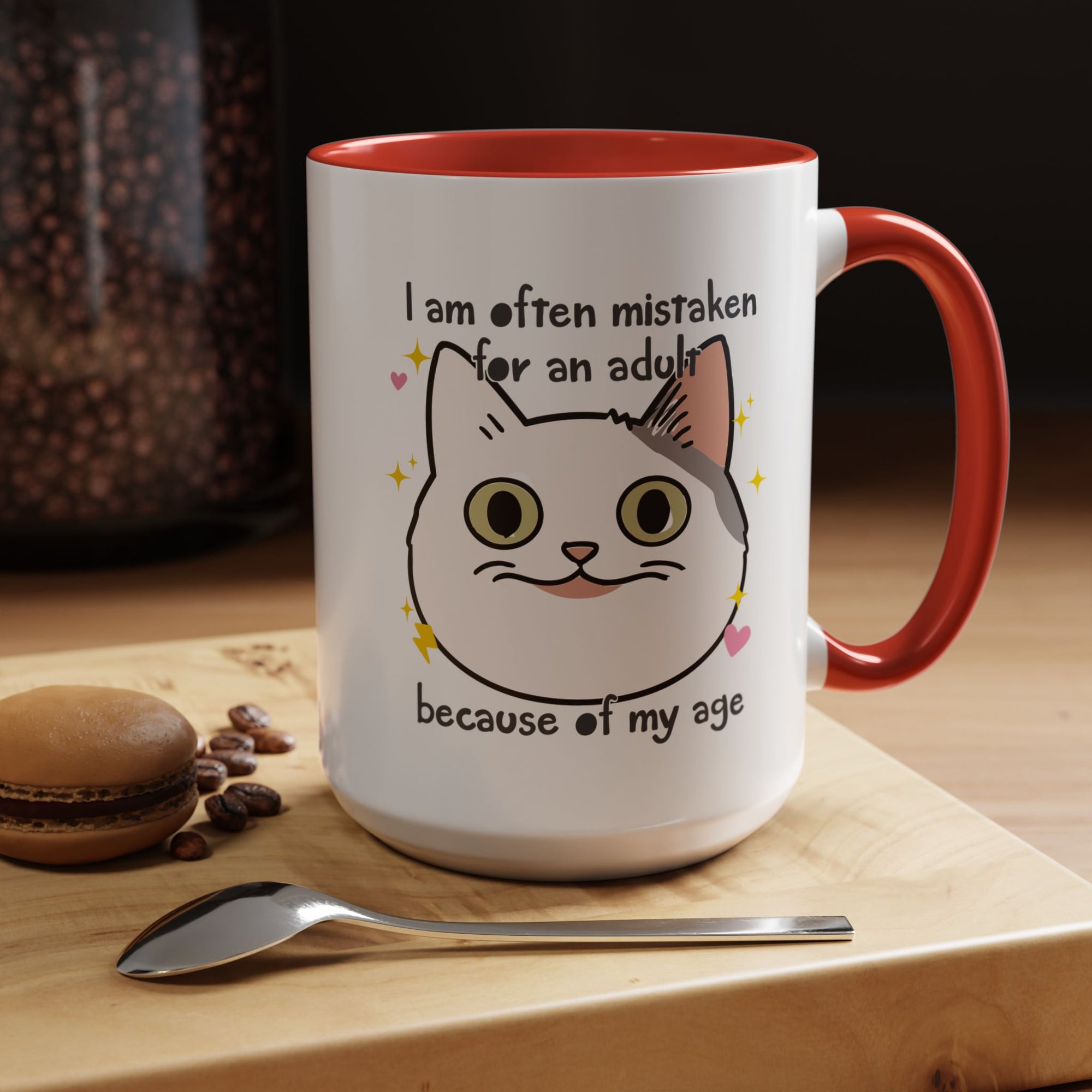 White ceramic mug with red handle and interior featuring a cute wide-eyed cat face, hearts, sparkles, and the phrase “I am often mistaken for an adult because of my age.”
