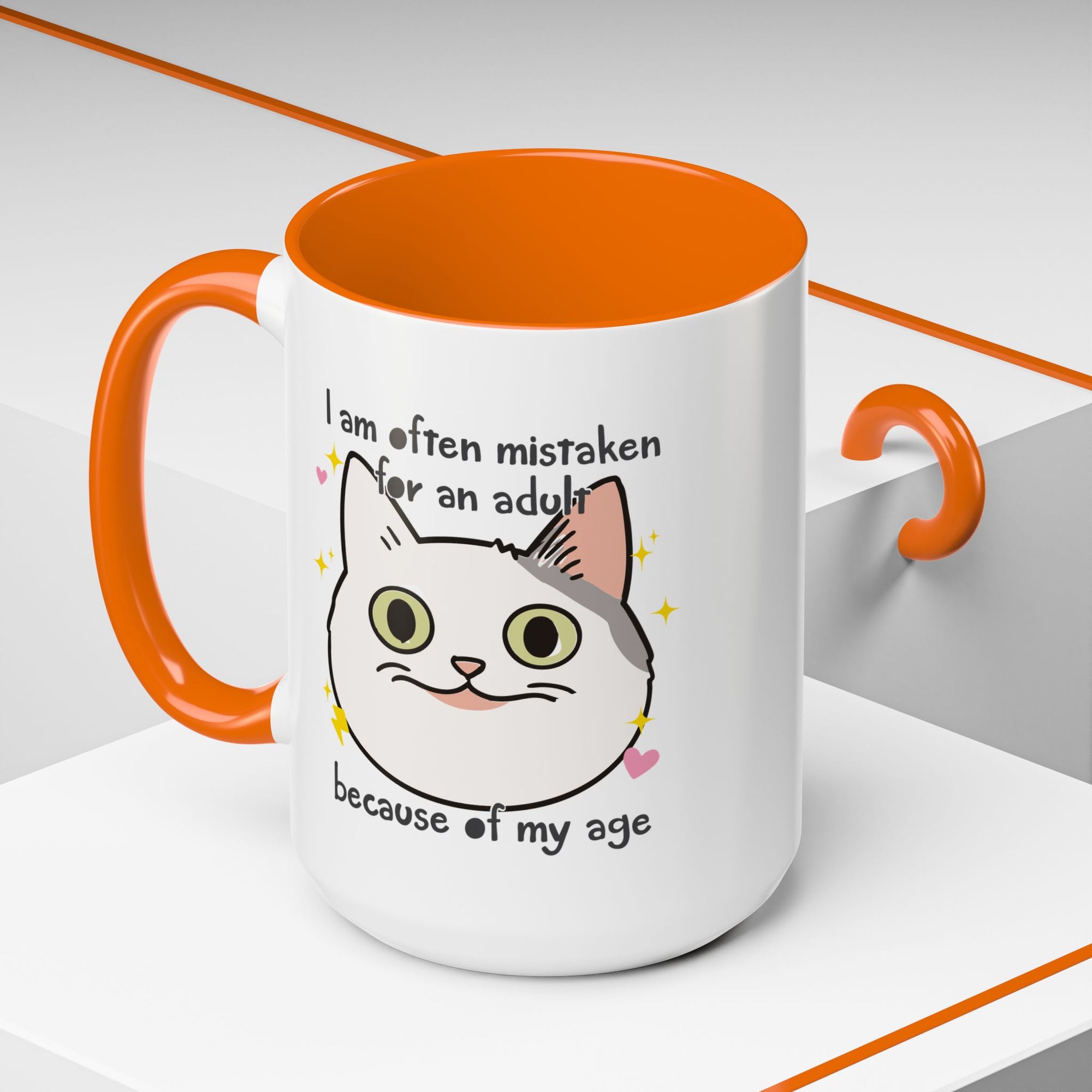 White ceramic mug with orange handle and interior featuring a cute wide-eyed cat face, hearts, sparkles, and the phrase “I am often mistaken for an adult because of my age.”
