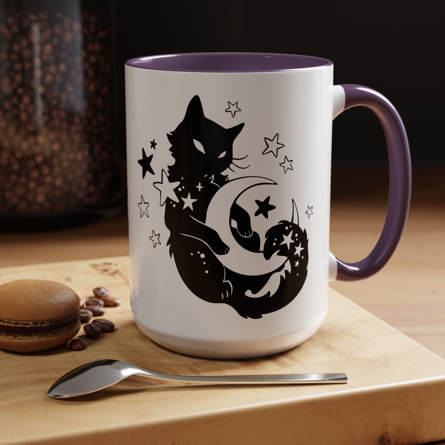 White mug with purple handle featuring black cat and moon design on a wooden surface with coffee beans and a macaron.