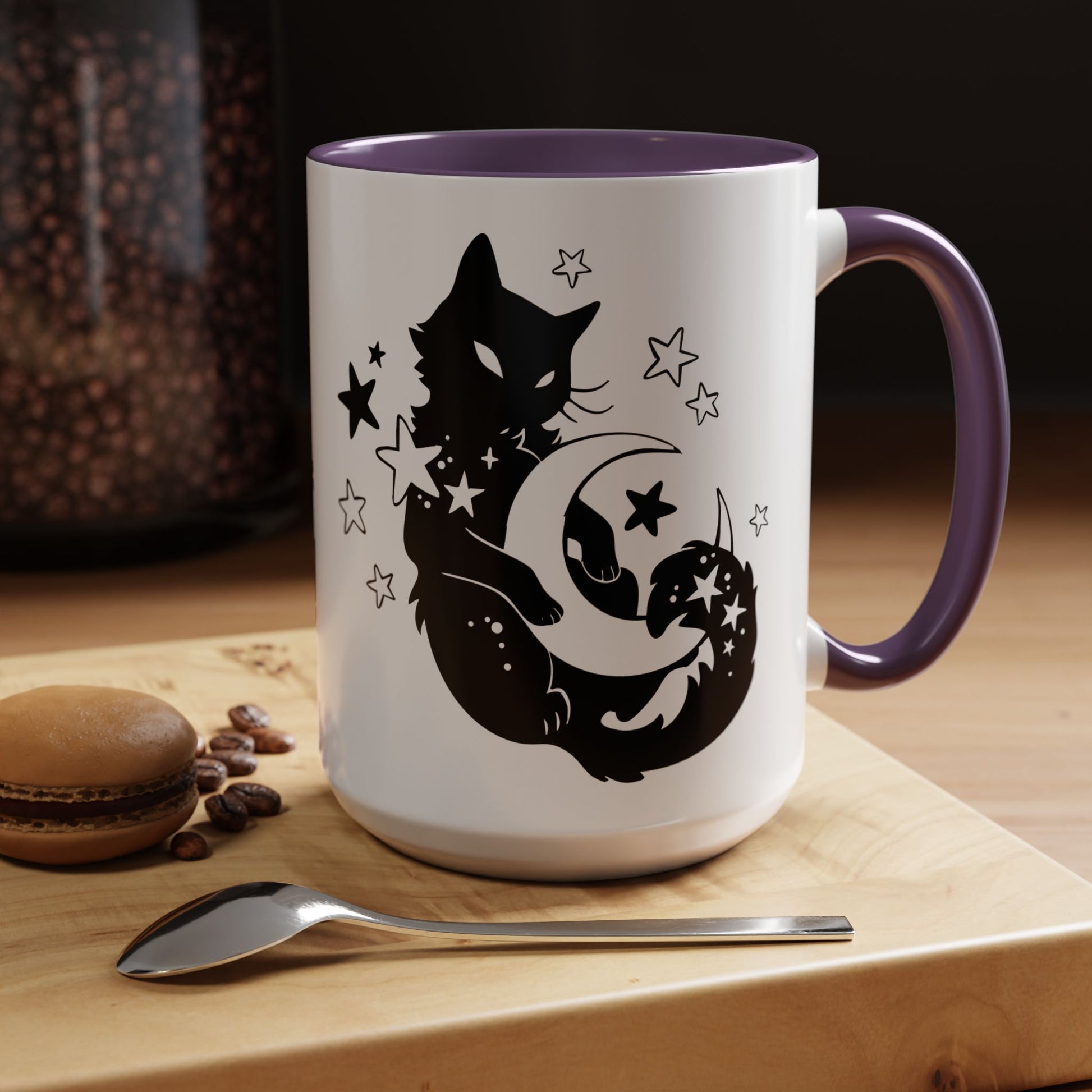 White mug with purple handle featuring black cat and moon design on a wooden surface with coffee beans and a macaron.