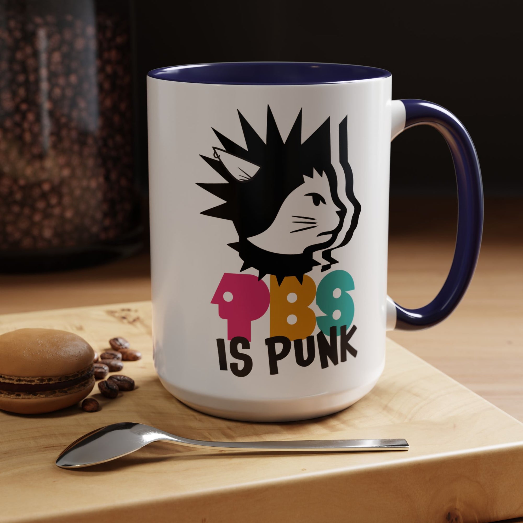 White mug with black interior and handle, featuring a cat graphic and 'PBS is Punk' text on a wooden surface with coffee beans and a macaron.