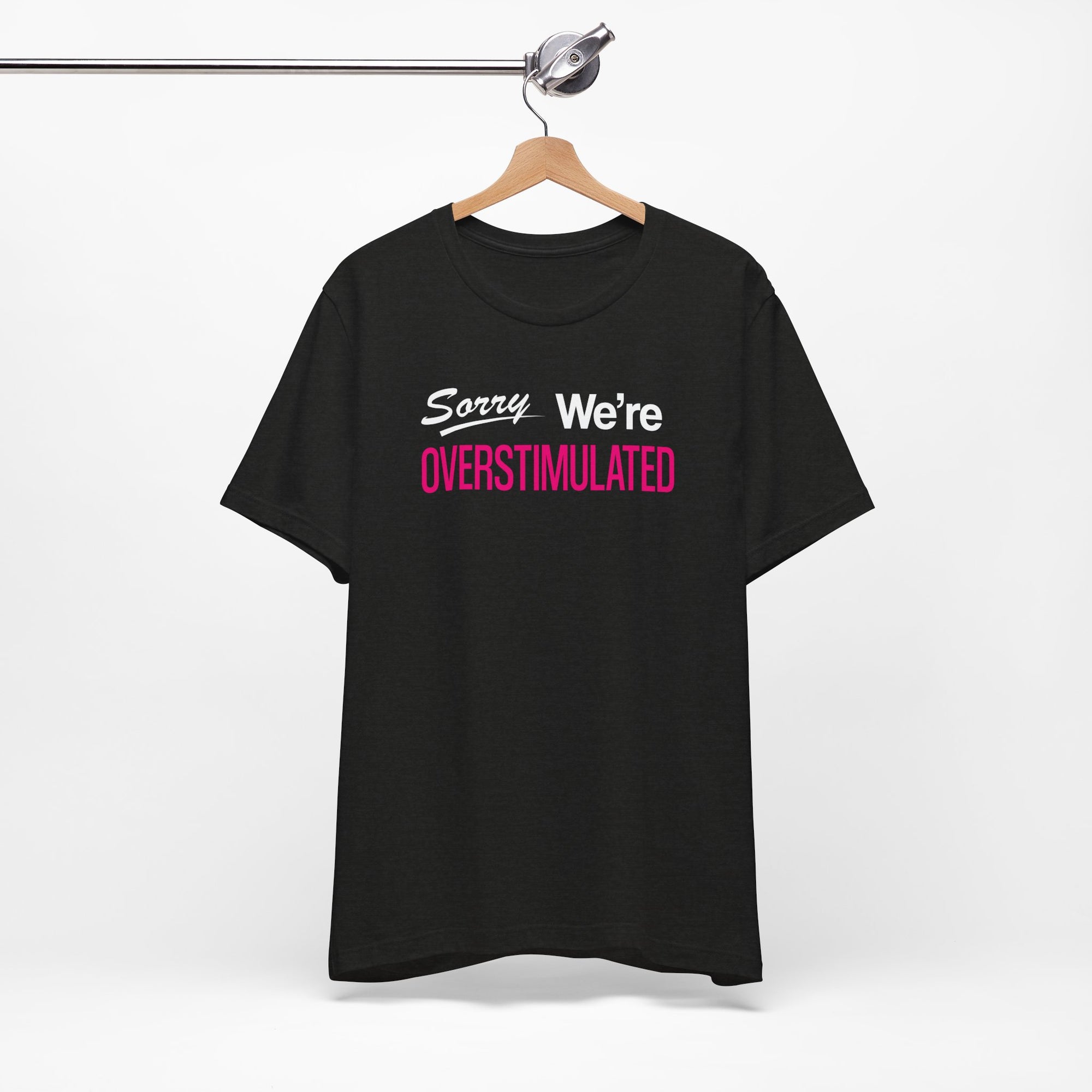 Black t-shirt featuring the phrase “Sorry We’re Overstimulated” in white and hot pink text.
