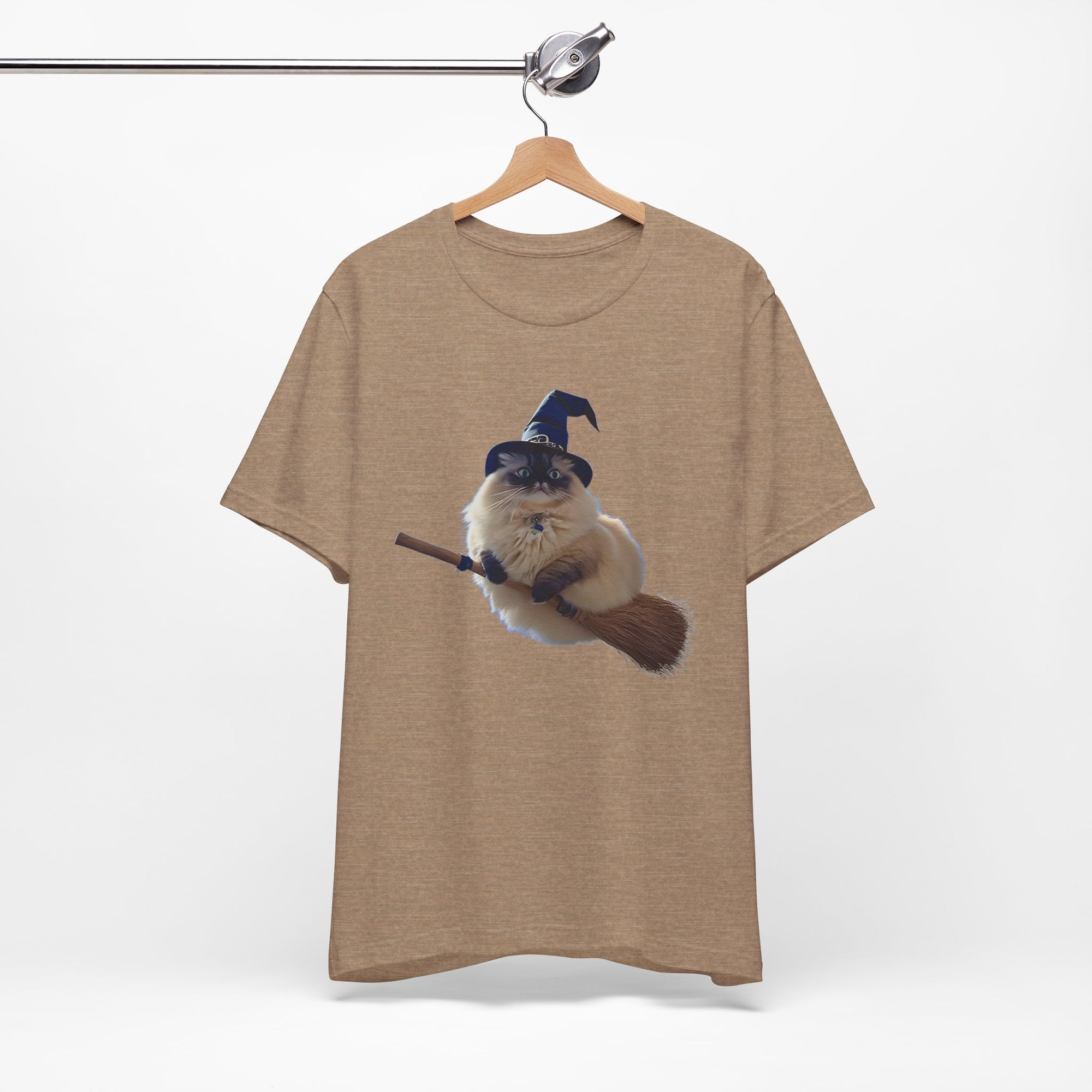 Beige t-shirt with a graphic of a cat in a wizard hat and on a broomstick, hanging on a wooden hanger against a white background.