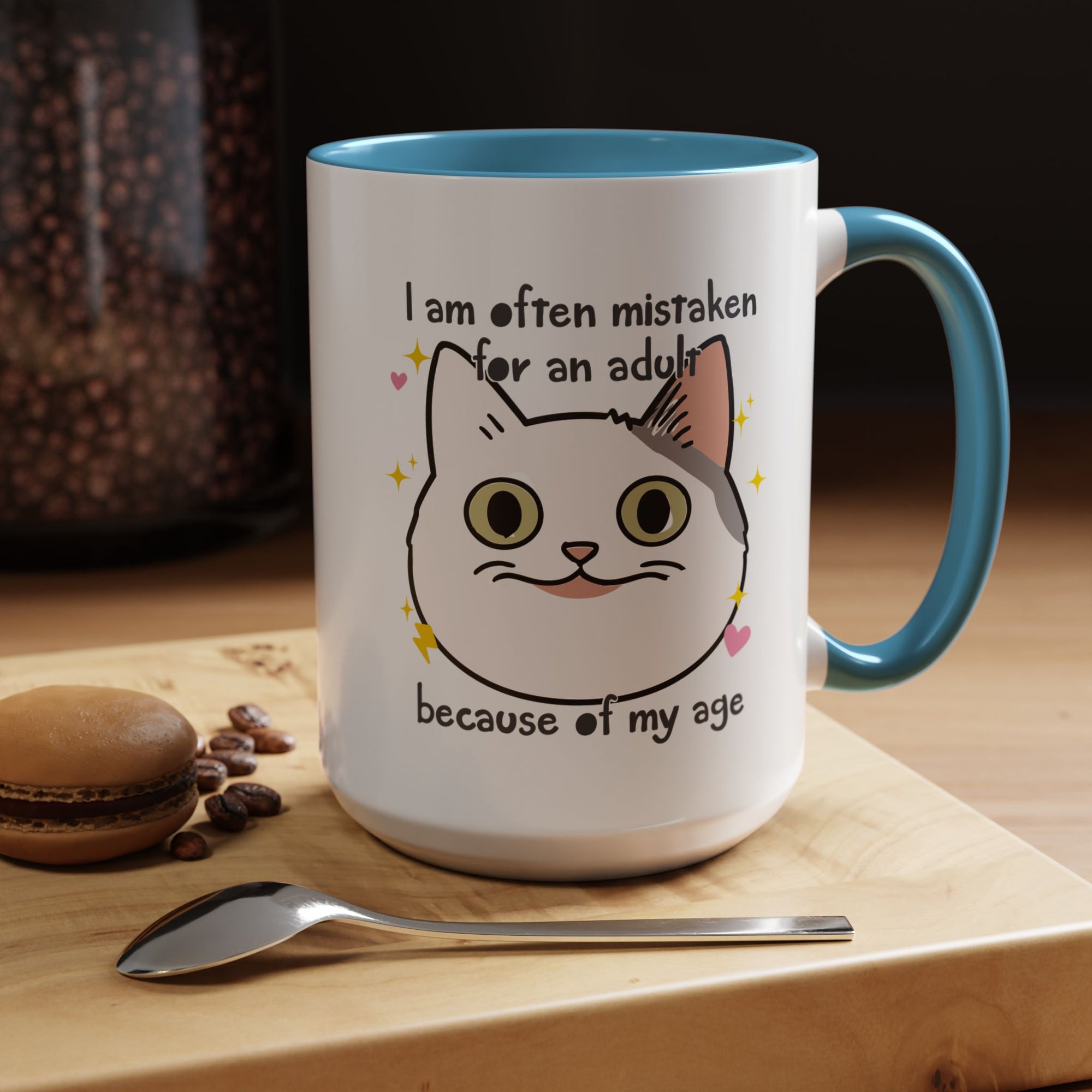 White ceramic mug with blue handle and interior featuring a cute wide-eyed cat face, hearts, sparkles, and the phrase “I am often mistaken for an adult because of my age.”
