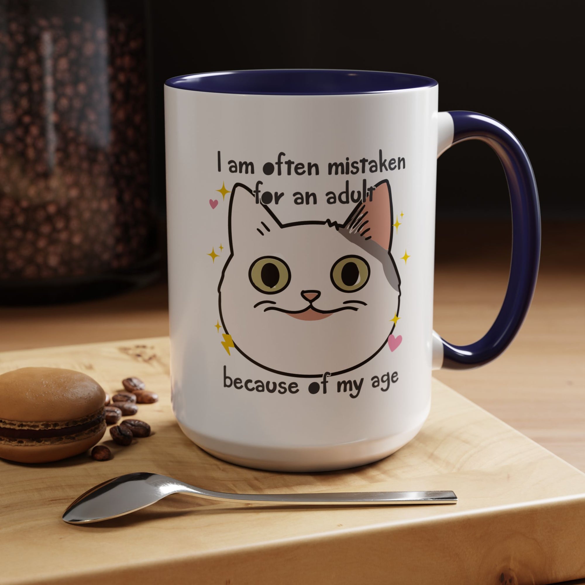 White ceramic mug with purple handle and interior featuring a cute wide-eyed cat face, hearts, sparkles, and the phrase “I am often mistaken for an adult because of my age.”
