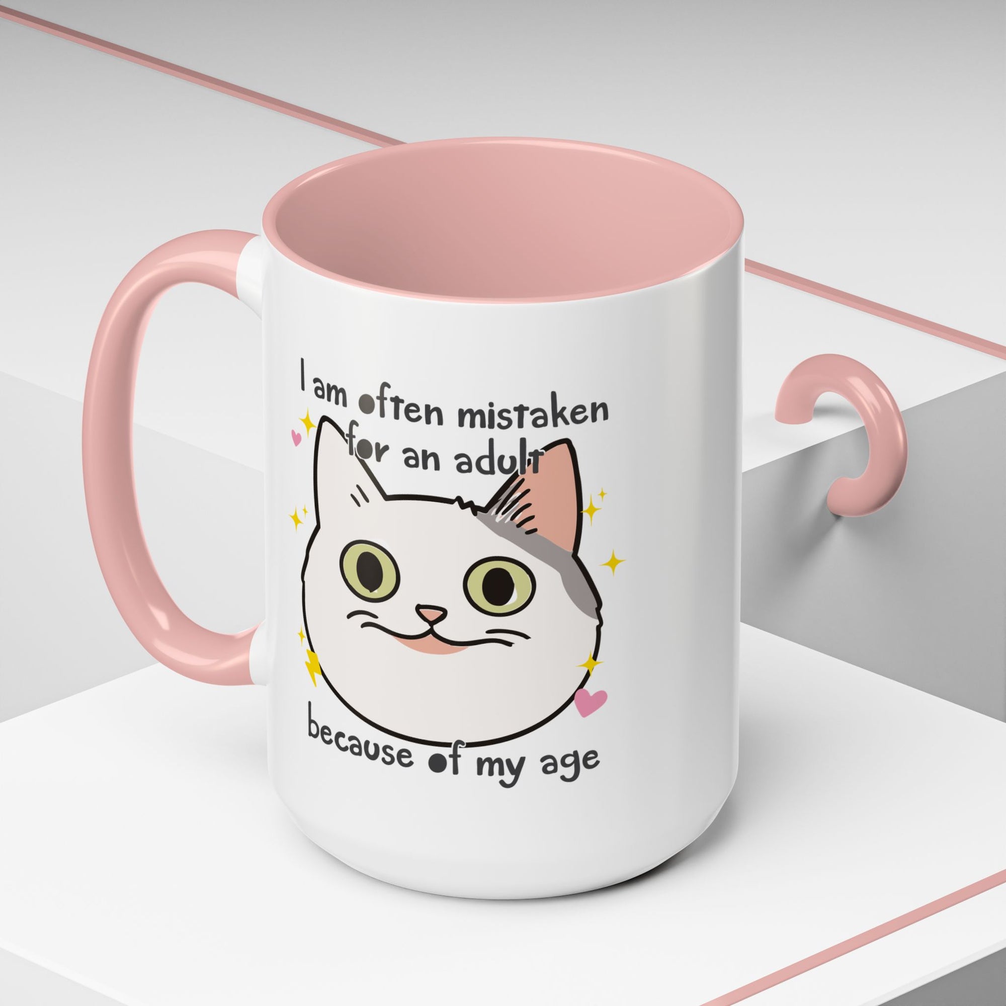 White ceramic mug with peach handle and interior featuring a cute wide-eyed cat face, hearts, sparkles, and the phrase “I am often mistaken for an adult because of my age.”
