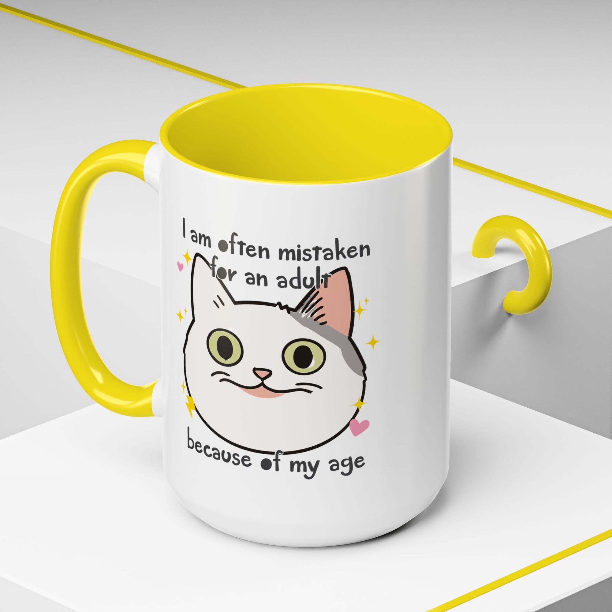 White ceramic mug with yellow handle and interior featuring a cute wide-eyed cat face, hearts, sparkles, and the phrase “I am often mistaken for an adult because of my age.”
