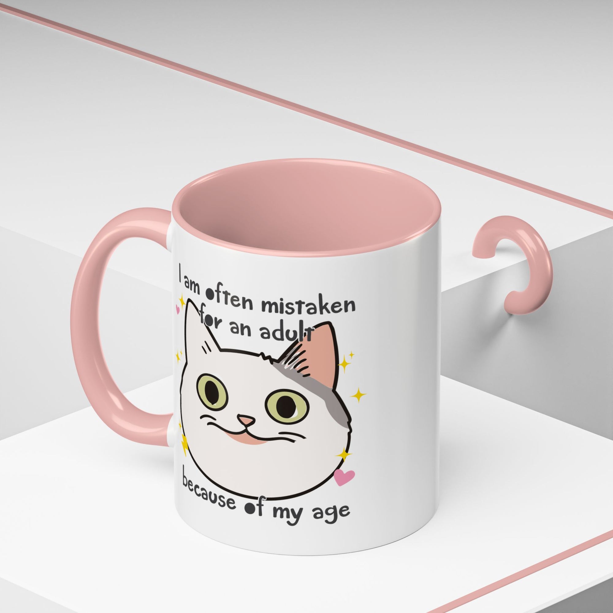 White ceramic mug with peach handle and interior featuring a cute wide-eyed cat face, hearts, sparkles, and the phrase “I am often mistaken for an adult because of my age.”
