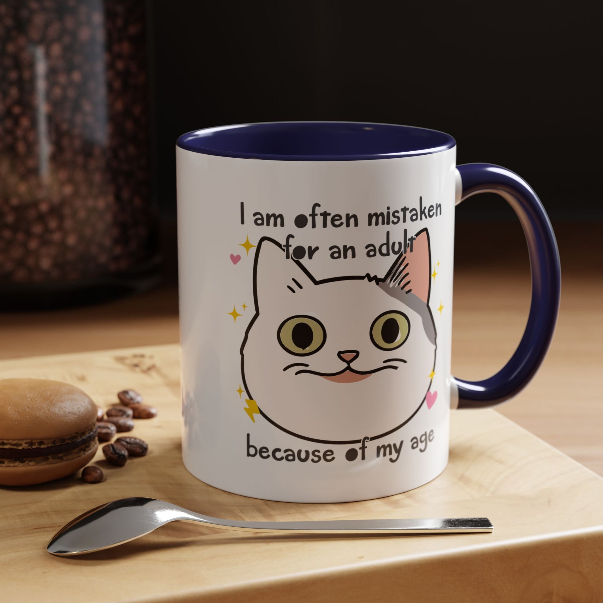 White ceramic mug with purple handle and interior featuring a cute wide-eyed cat face, hearts, sparkles, and the phrase “I am often mistaken for an adult because of my age.”
