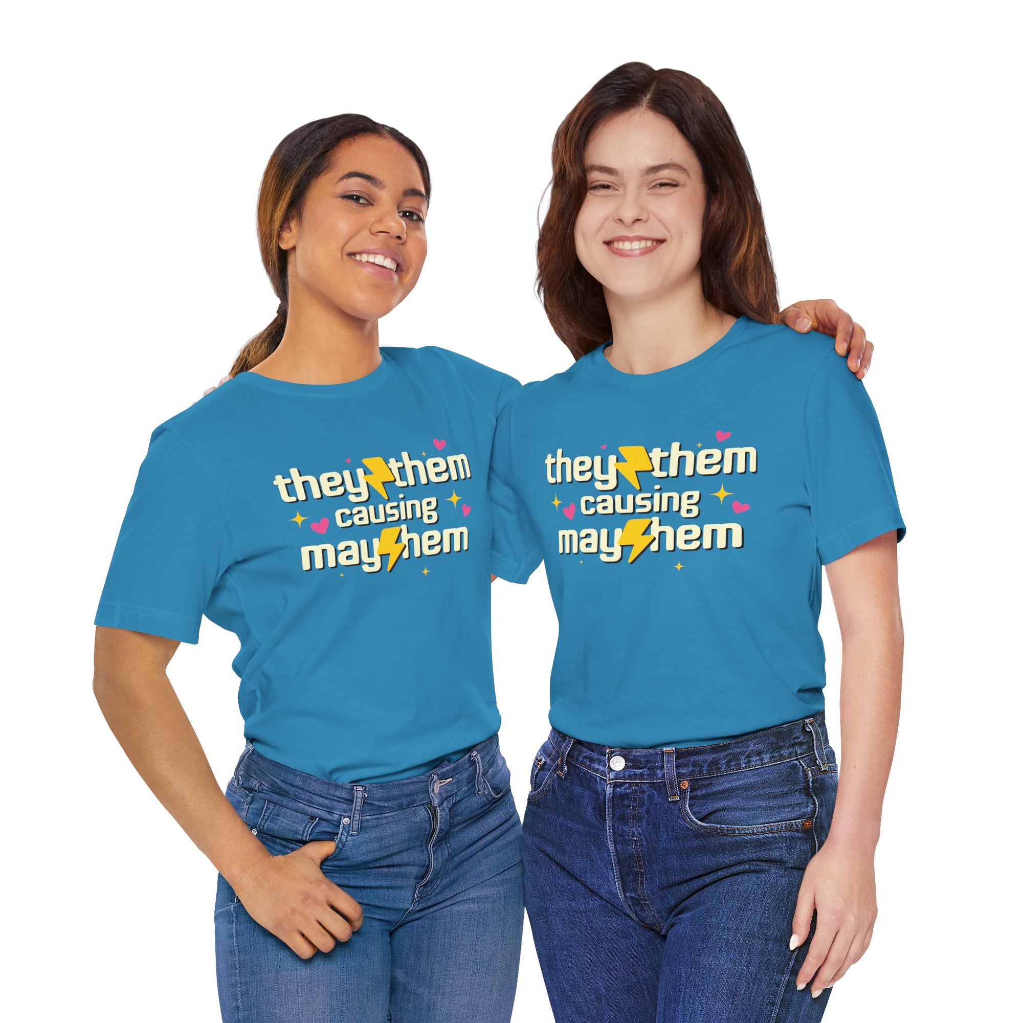 Two people wearing blue shirts featuring the phrase “They/Them Causing Mayhem” in retro-style lettering with yellow lightning bolts, pink hearts, and sparkle accents.
