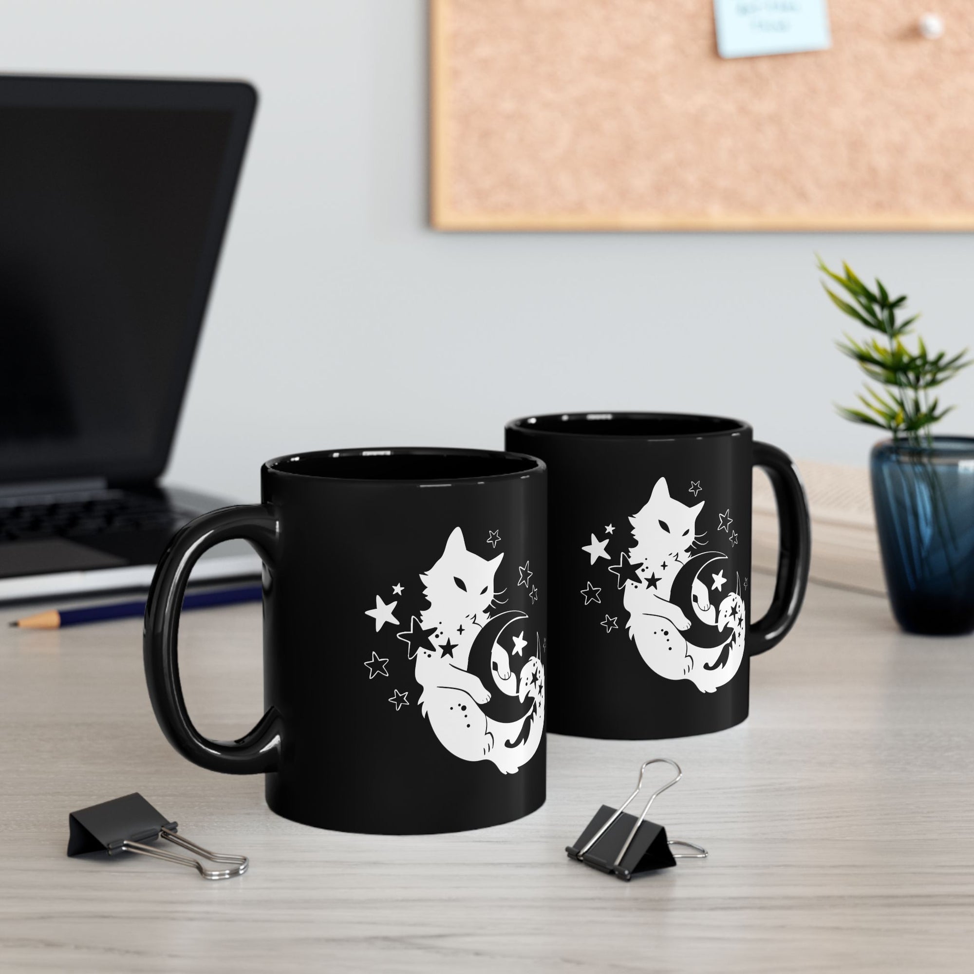 Two black mugs with cat designs on a desk with office items.
