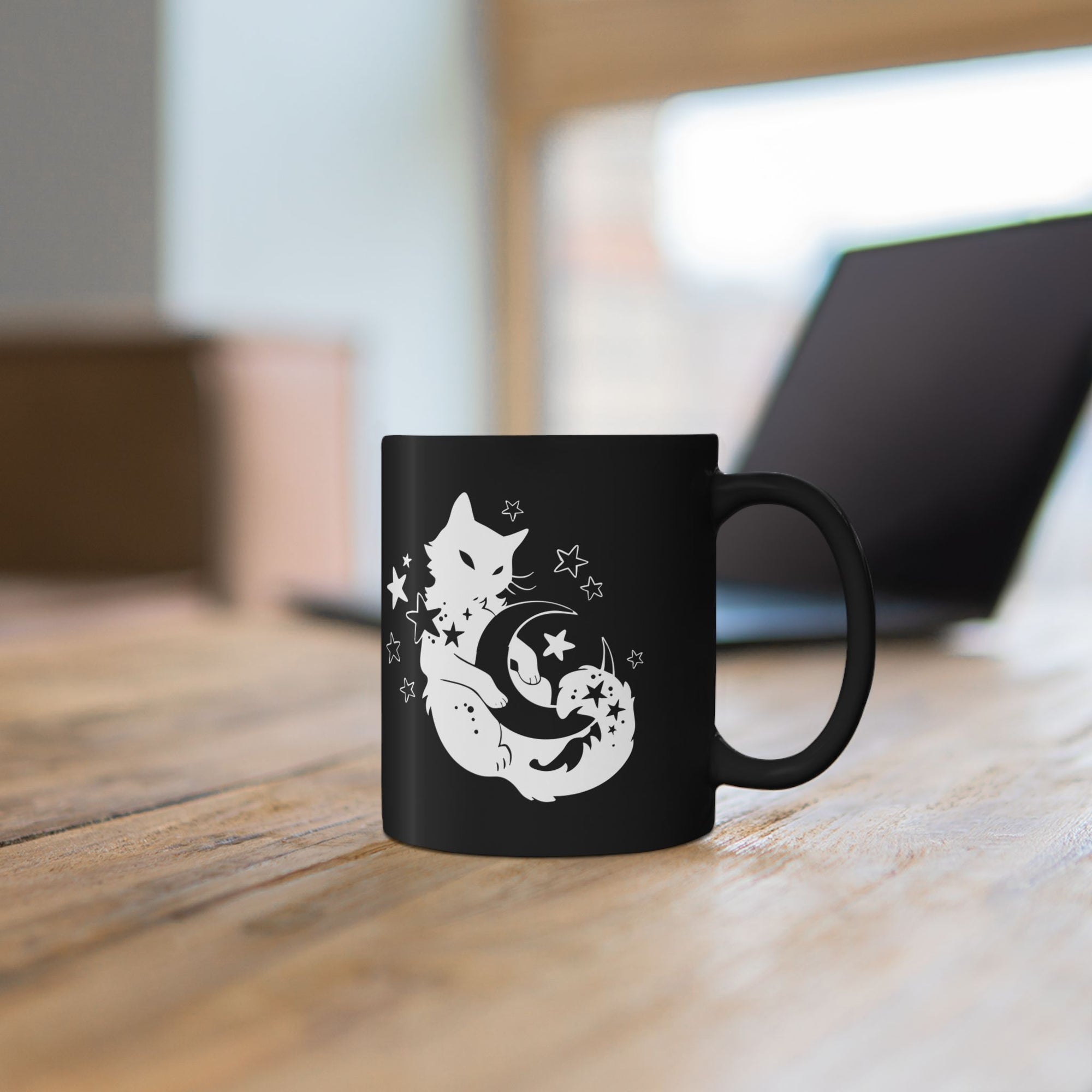 Black mug with a white cat and moon design on a wooden surface.