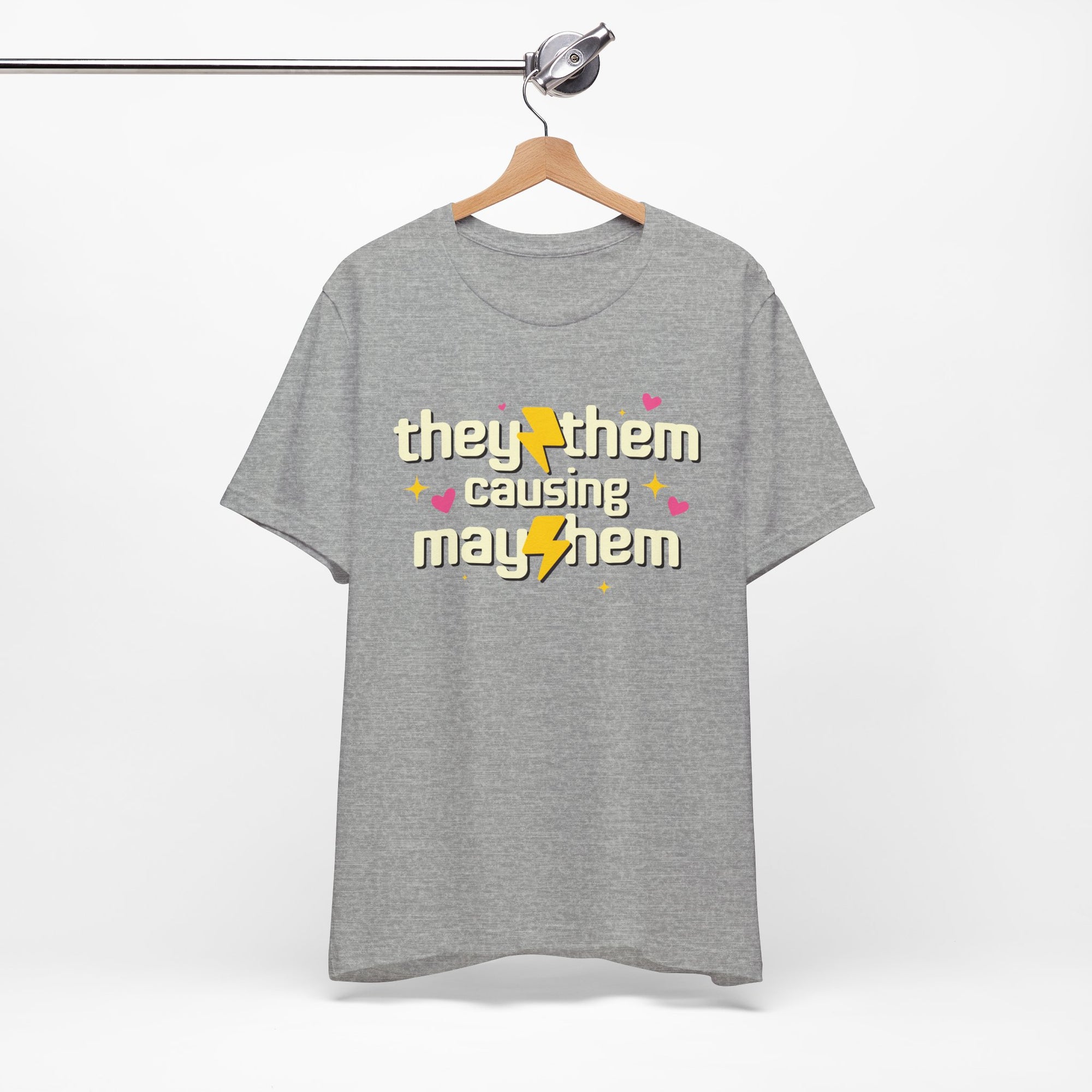 Gray t-shirt with 'they/them causing mayhem' text on a hanger against a white background