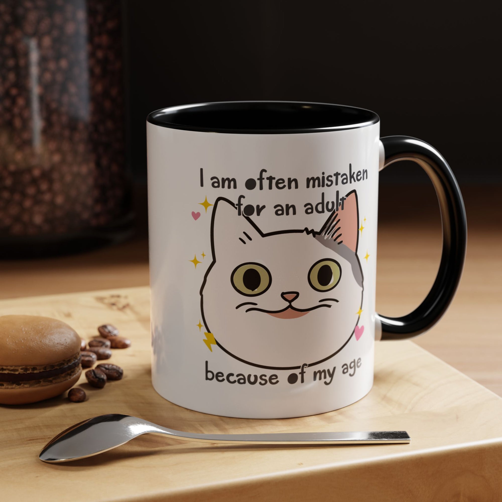 White ceramic mug with black handle and interior featuring a cute wide-eyed cat face, hearts, sparkles, and the phrase “I am often mistaken for an adult because of my age.”
