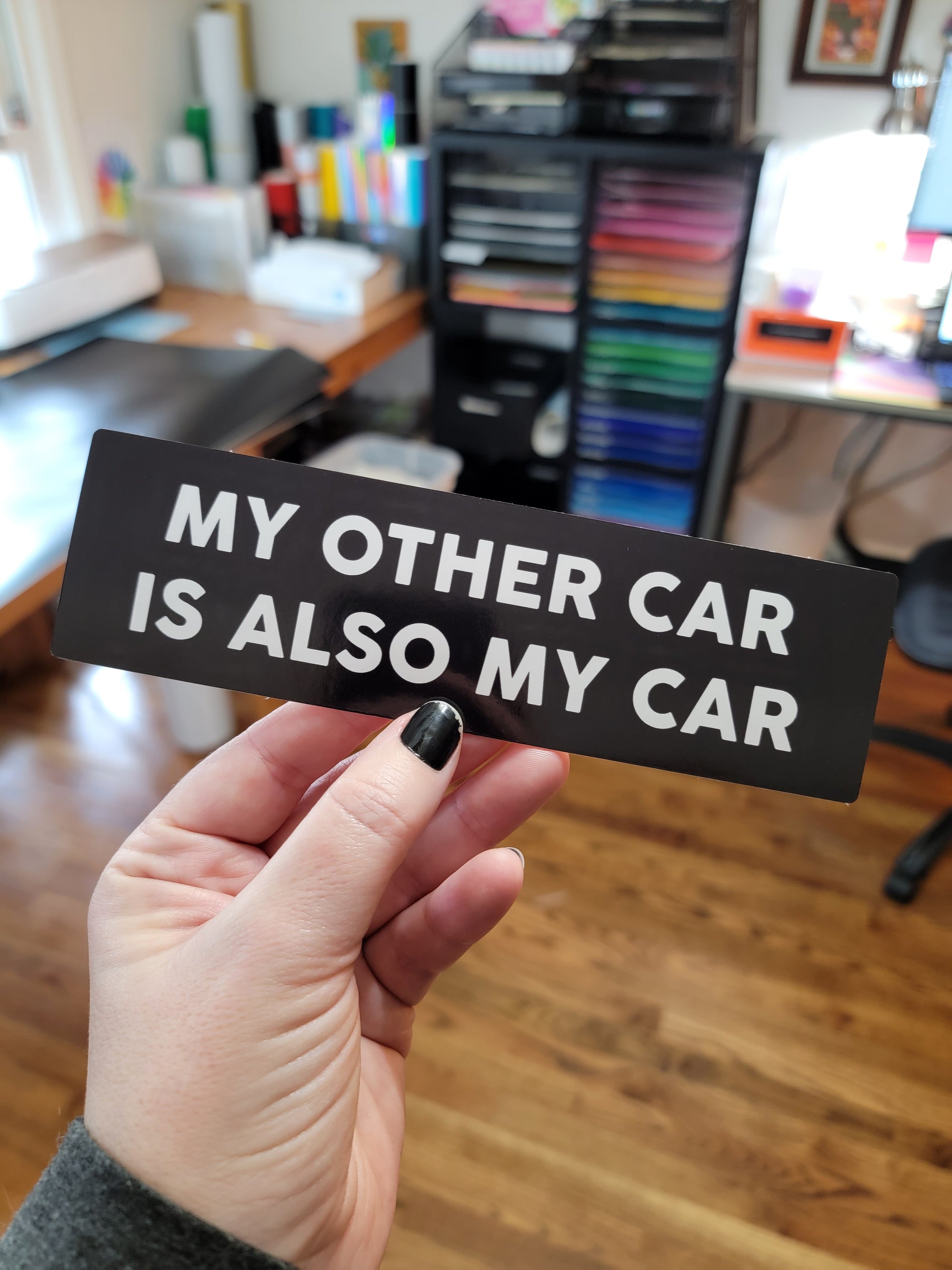 Rectangular black bumper sticker with bold white text that says “My Other Car Is Also My Car.”
