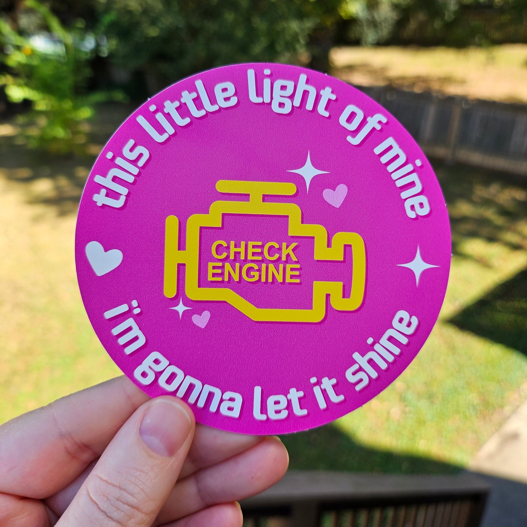 round vinyl stickers, on pink featuring a yellow check engine light icon with the phrase “this little light of mine i’m gonna let it shine.”
