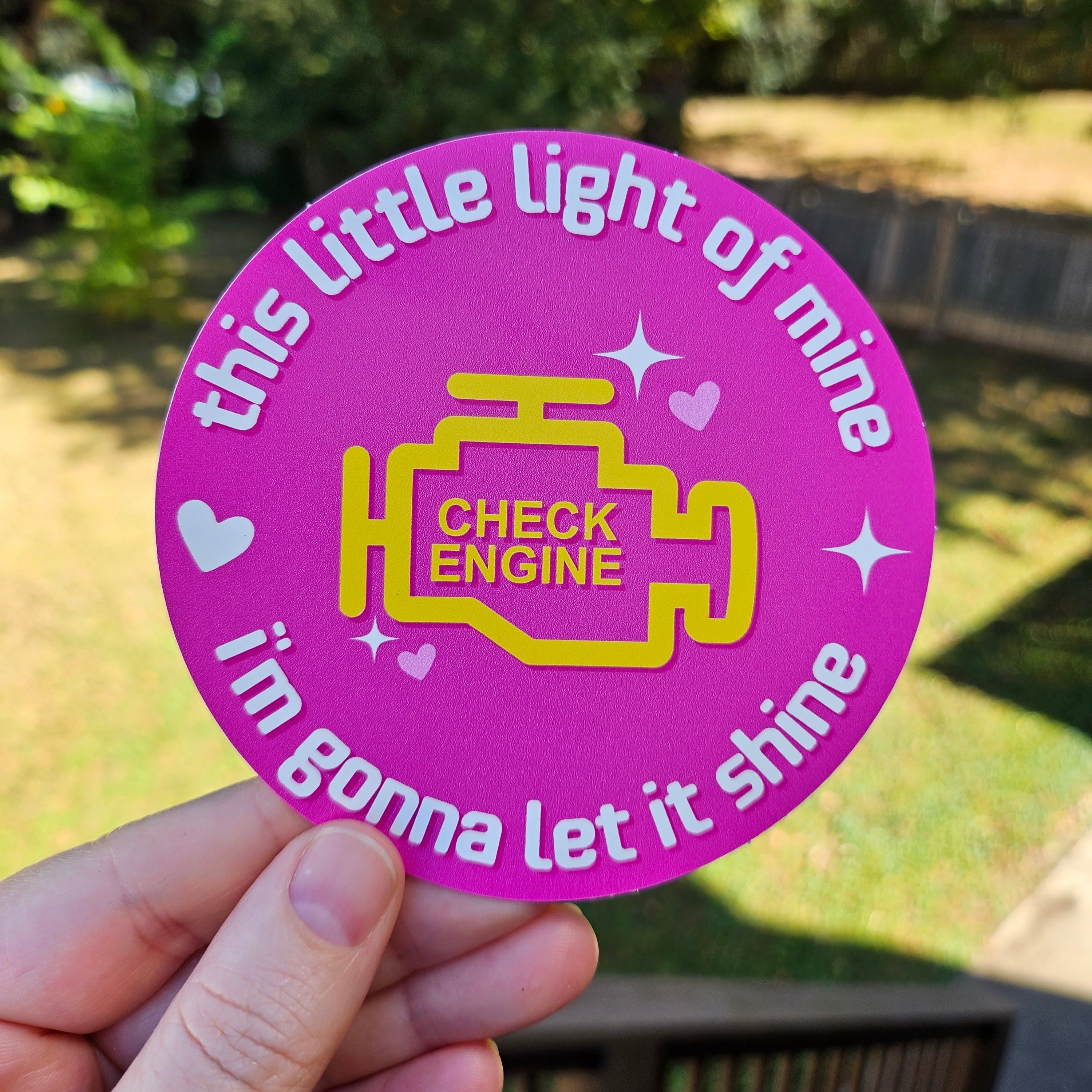 round vinyl stickers, on pink featuring a yellow check engine light icon with the phrase “this little light of mine i’m gonna let it shine.”

