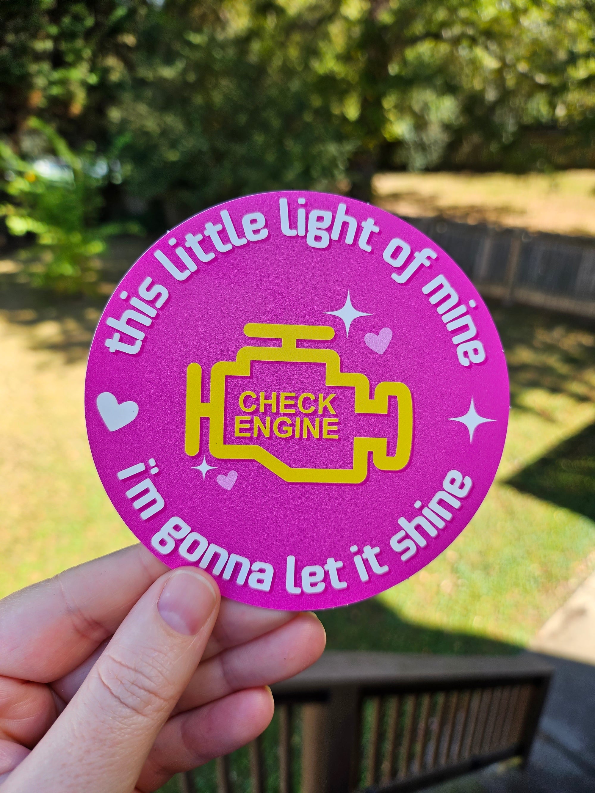 A hand holds a round pink sticker with a yellow engine graphic and the text 'this little light of mine i'm gonna let it shine' in a sunny outdoor setting.