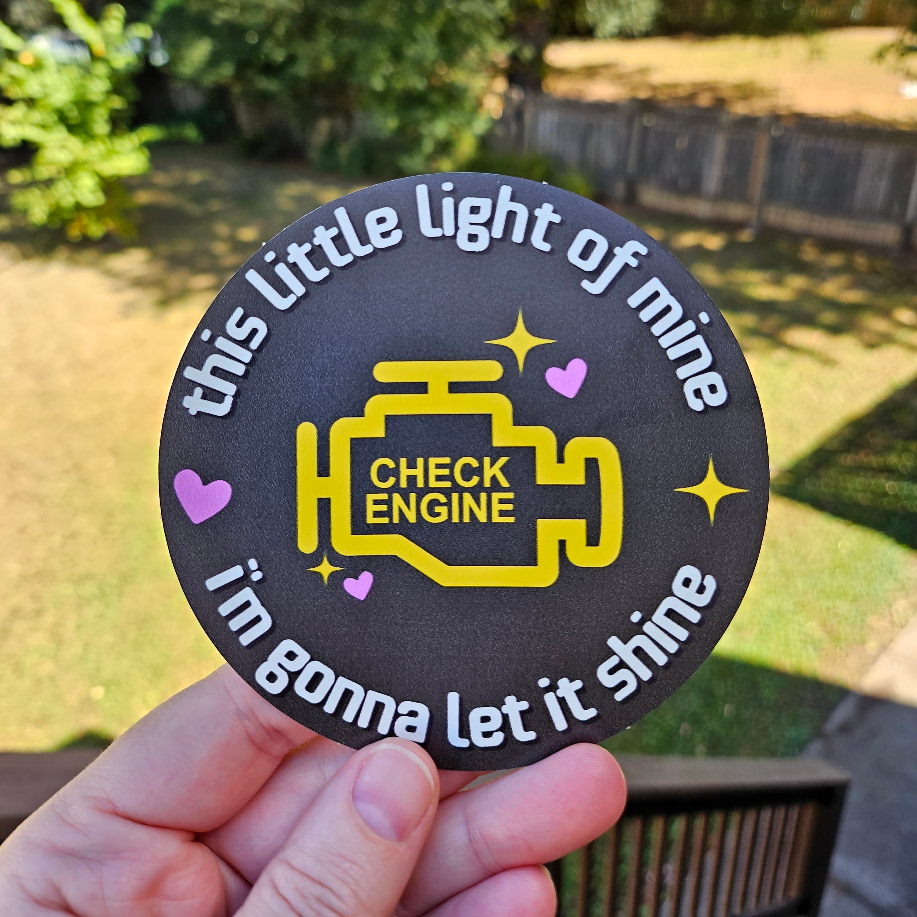 round vinyl sticker on dark gray featuring a yellow check engine light icon with the phrase “this little light of mine i’m gonna let it shine.”
