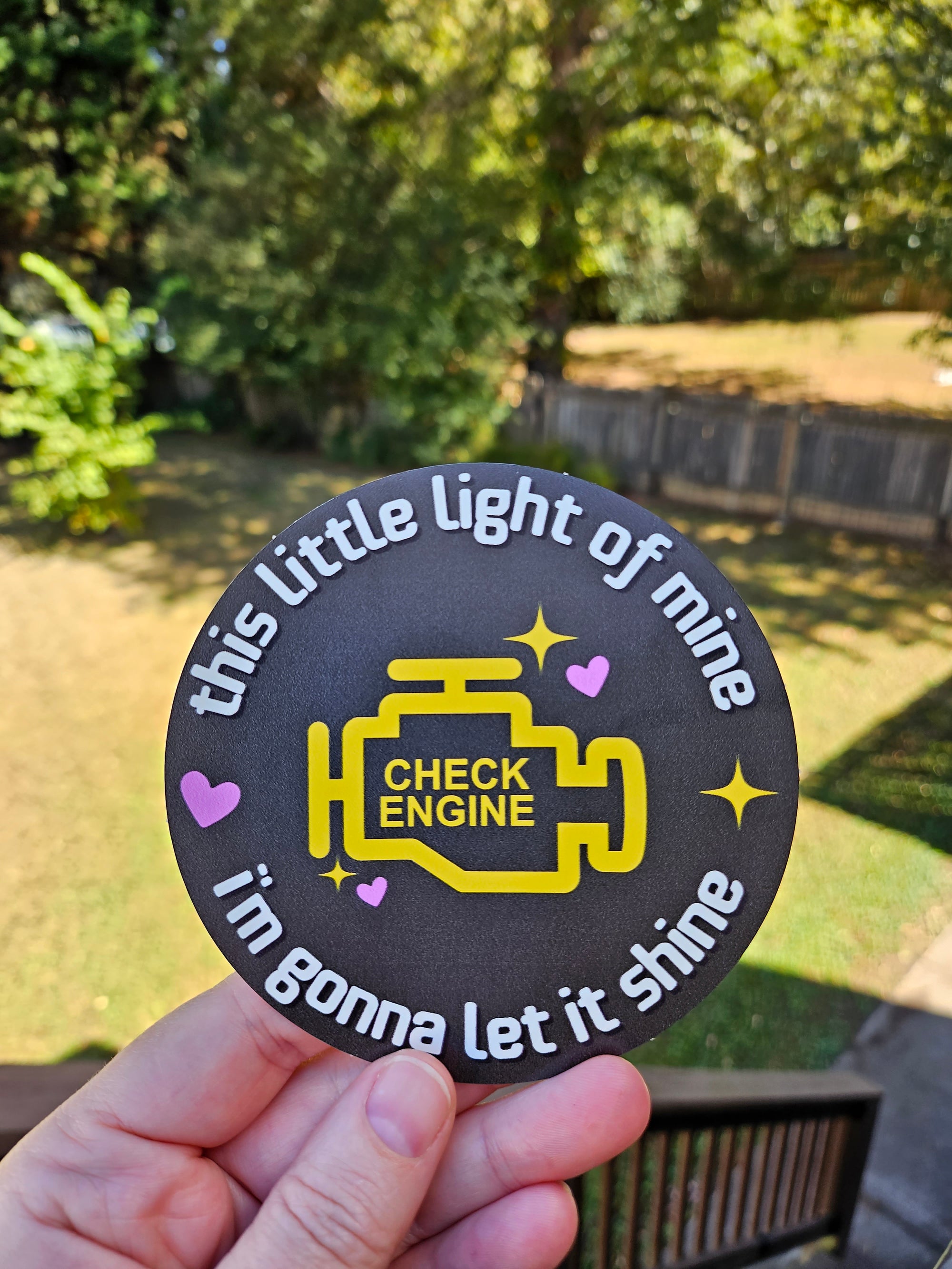 A hand holding a round black sticker outdoors. The sticker features a yellow engine graphic with the words 'CHECK ENGINE' and white text reading 'this little light of mine this big flame of love!'.
