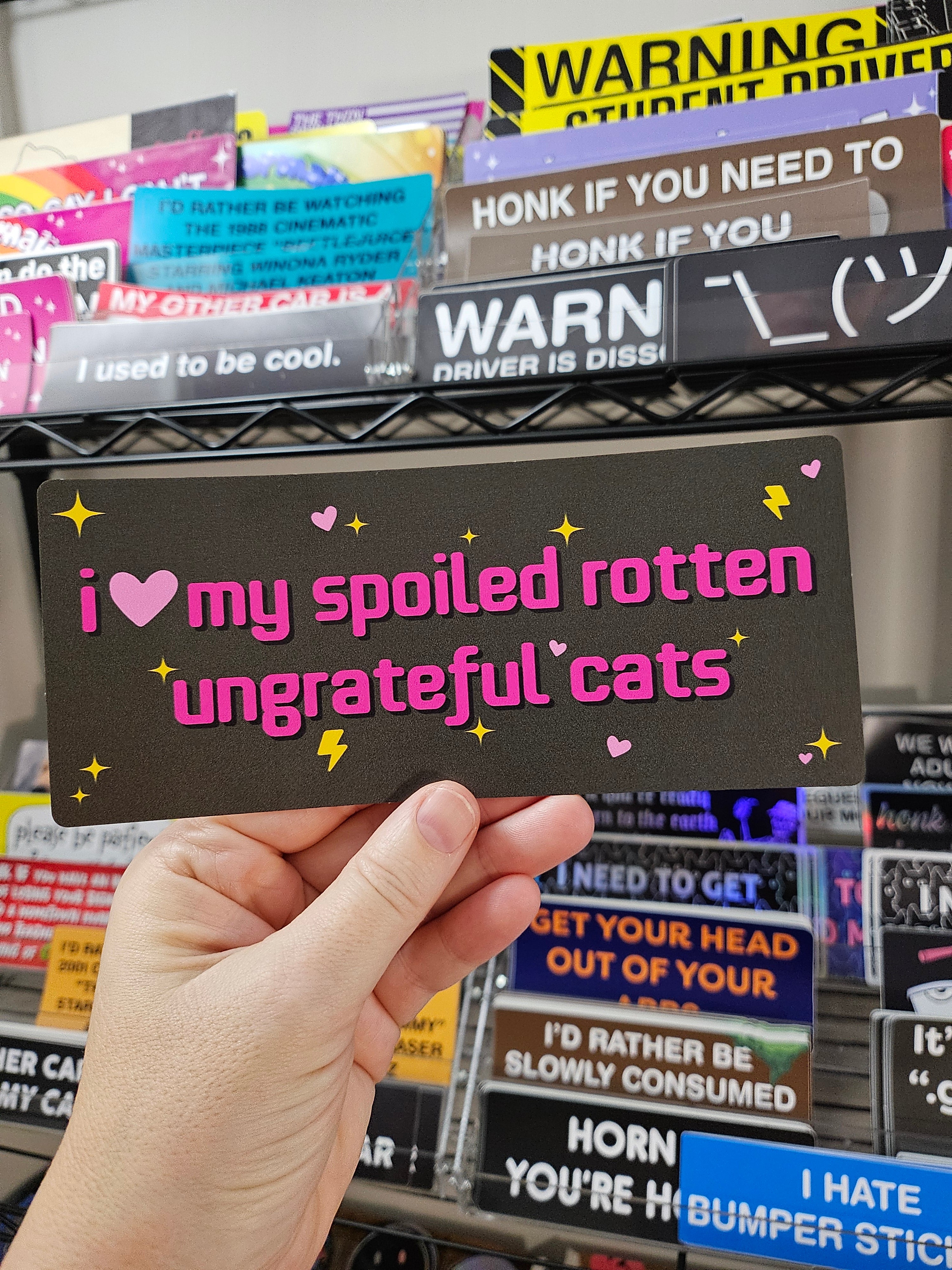 rectangular bumper stickers that say “I love my spoiled rotten ungrateful cats,” shown in dark gray color with hearts, sparkles, and lightning bolts.

