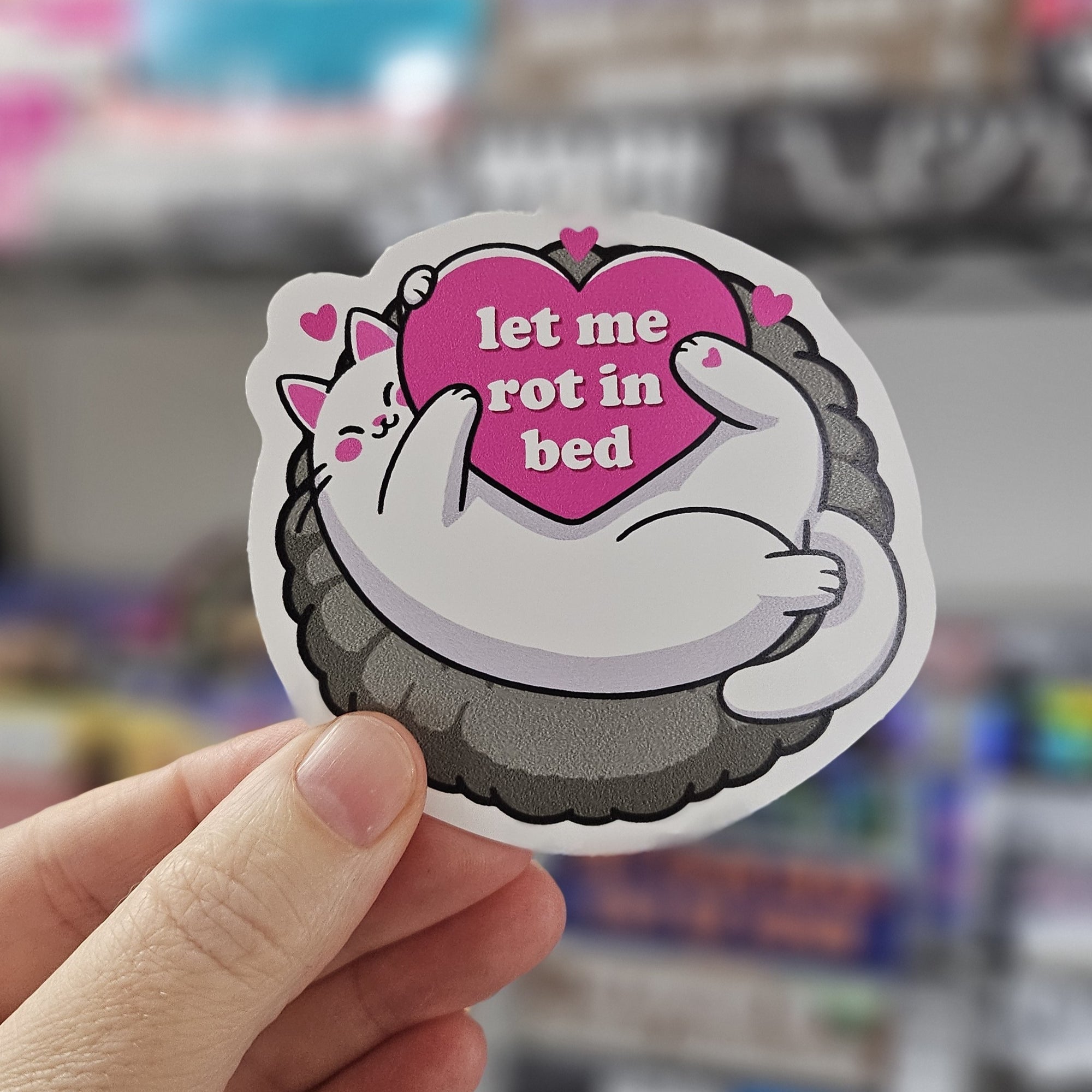round cozy cat vinyl stickers featuring a cat curled in a fluffy bed holding a pink heart, with “Let Me Rot in Bed” 
