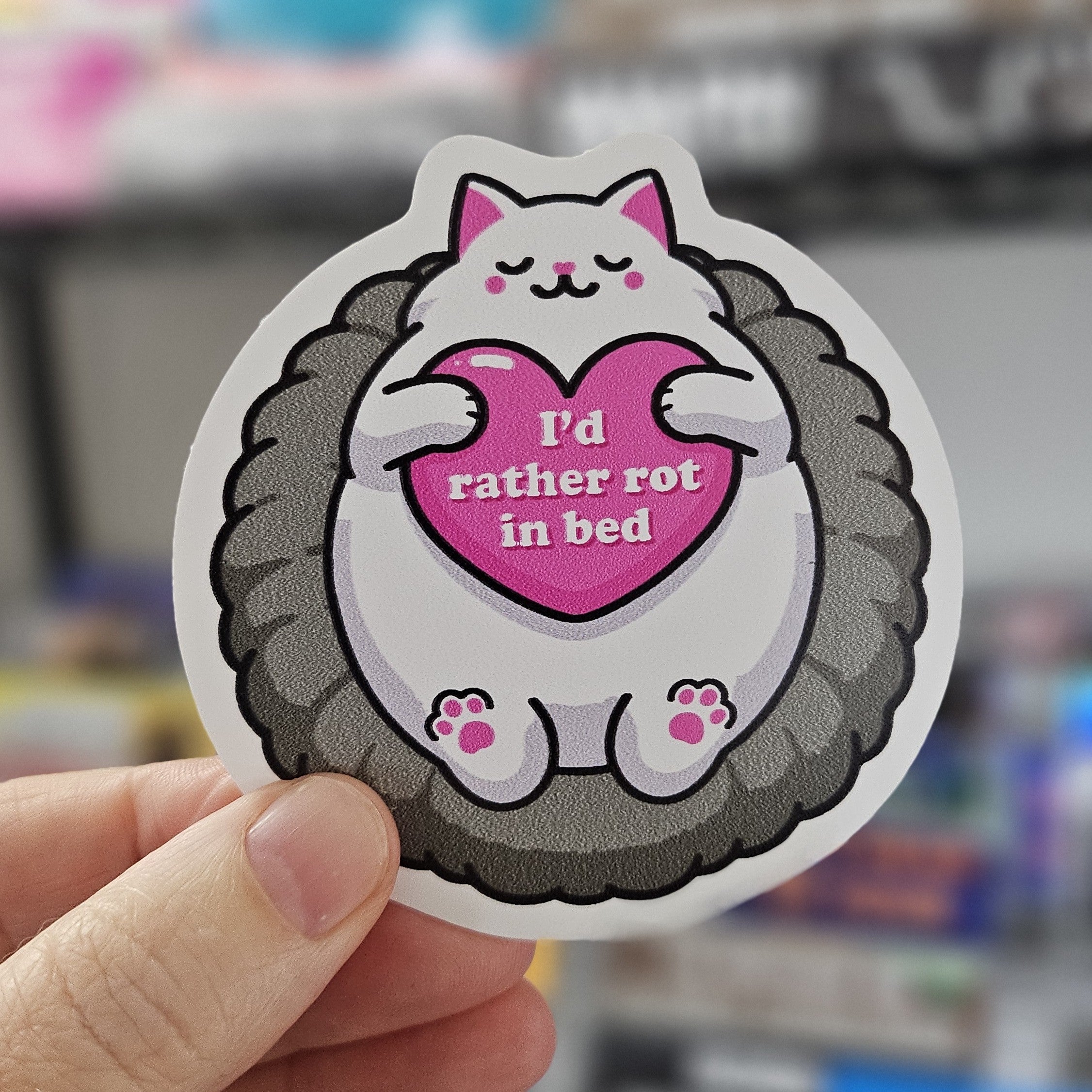 round cozy cat vinyl stickers featuring a cat curled in a fluffy bed holding a pink heart, with “I’d Rather Rot in Bed.”

