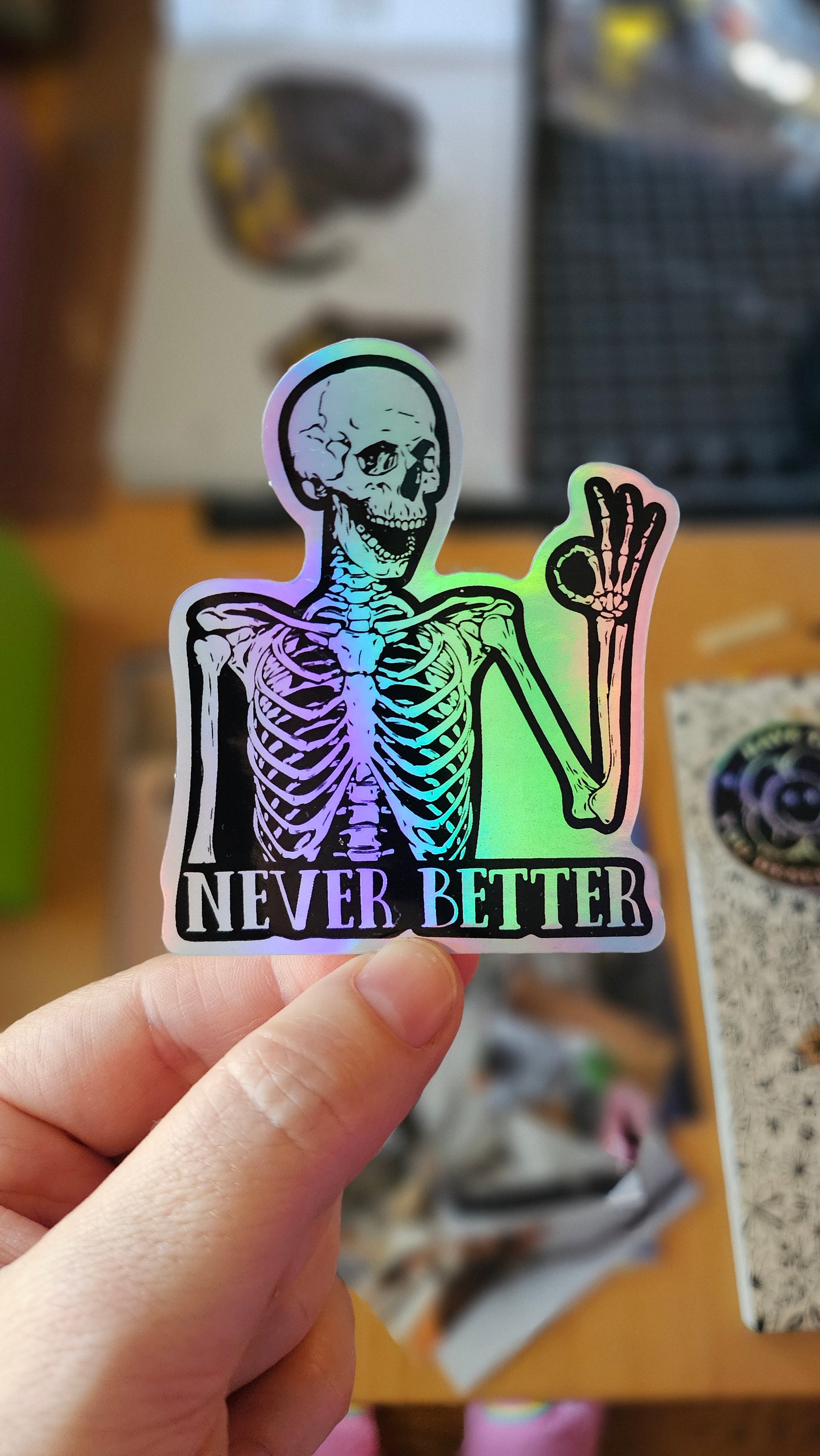 Skeleton sticker with the words “Never Better,” shown in white vinyl and holographic vinyl, featuring a smiling skeleton making an OK hand sign.