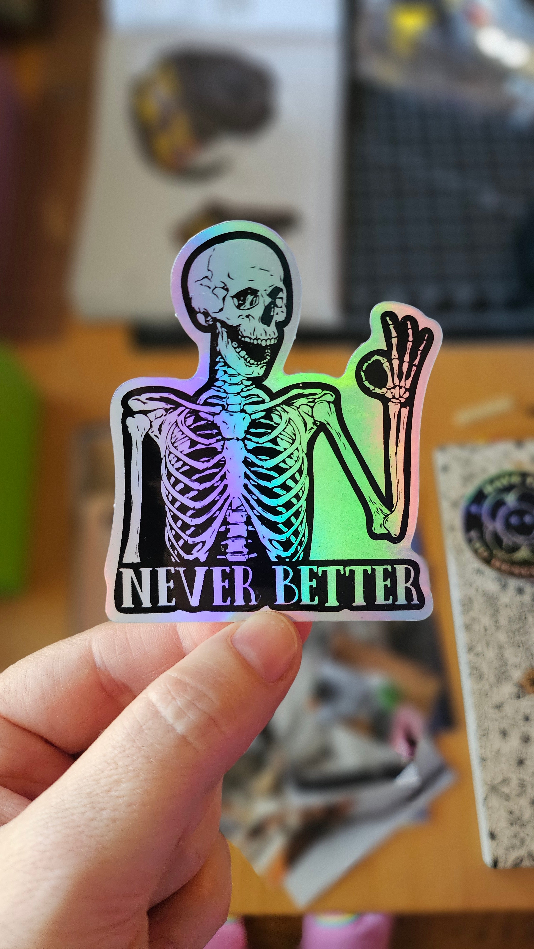 Skeleton sticker with the words “Never Better,” shown in white vinyl and holographic vinyl, featuring a smiling skeleton making an OK hand sign.