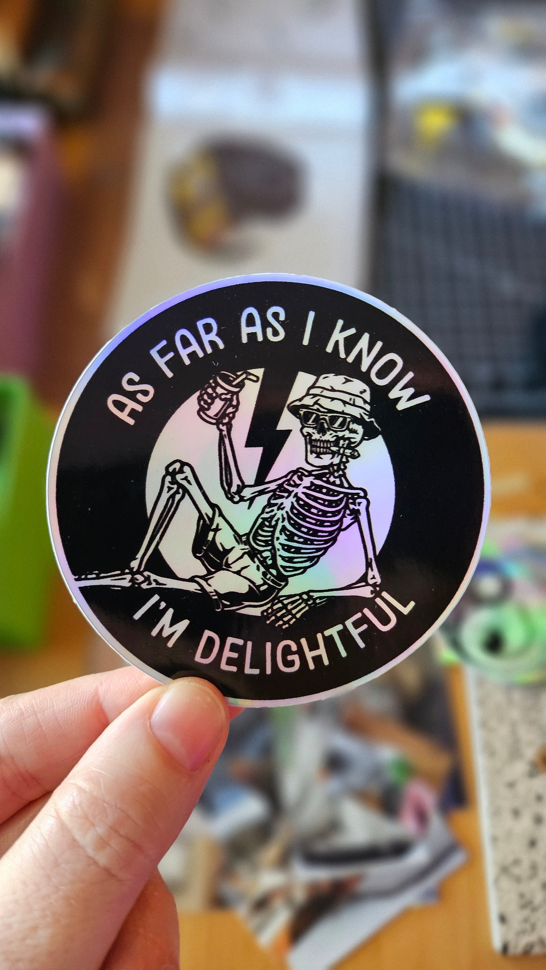 A circular sticker featuring a skeleton wearing a hat and sunglasses, with the text 'AS FAR AS I KNOW I'M DELIGHTFUL' and 'AS FAR AS I KNOW' around the border. The sticker is held by a hand against a blurred background.