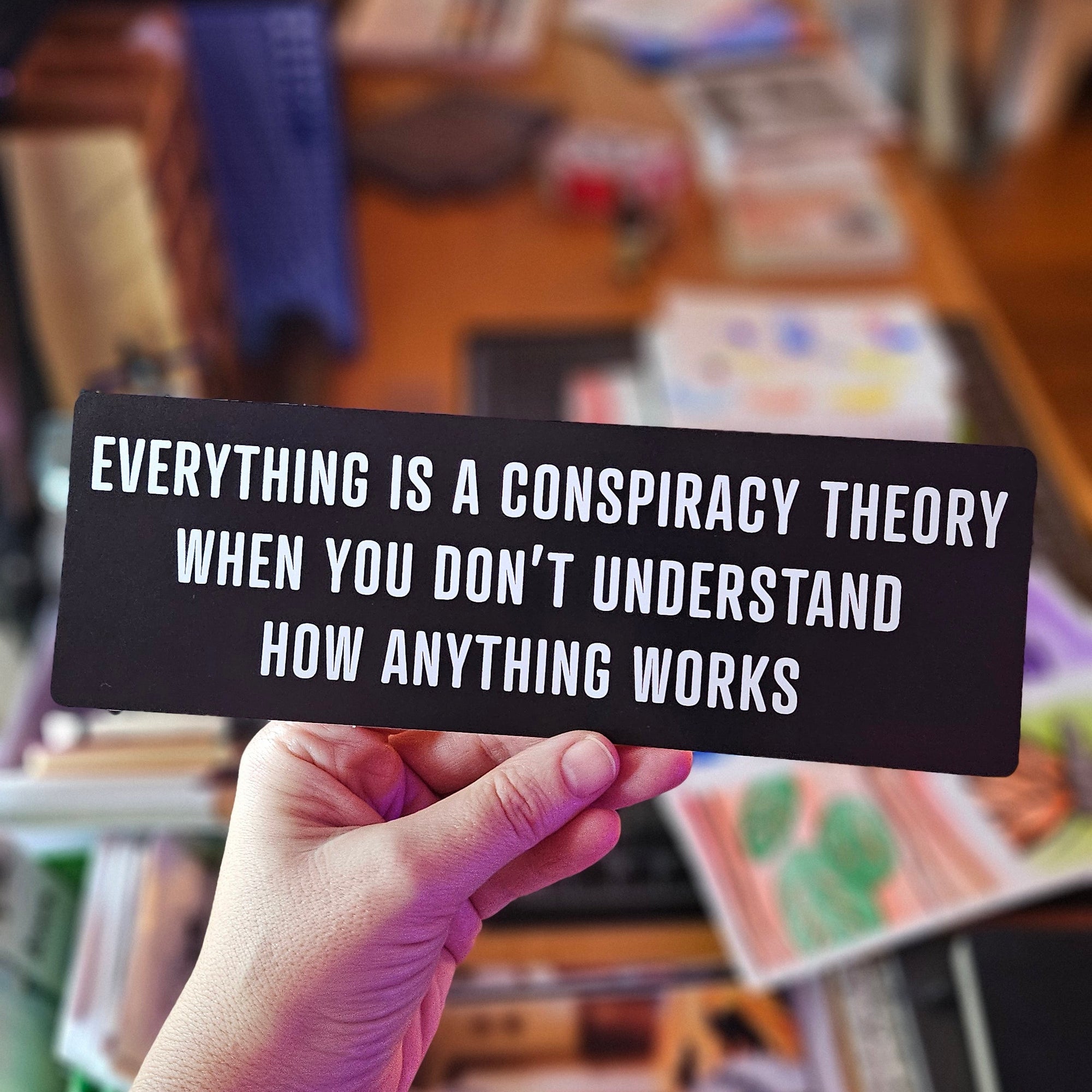 Rectangular black vinyl sticker with white text that says “Everything Is a Conspiracy Theory When You Don’t Understand How Anything Works.”
