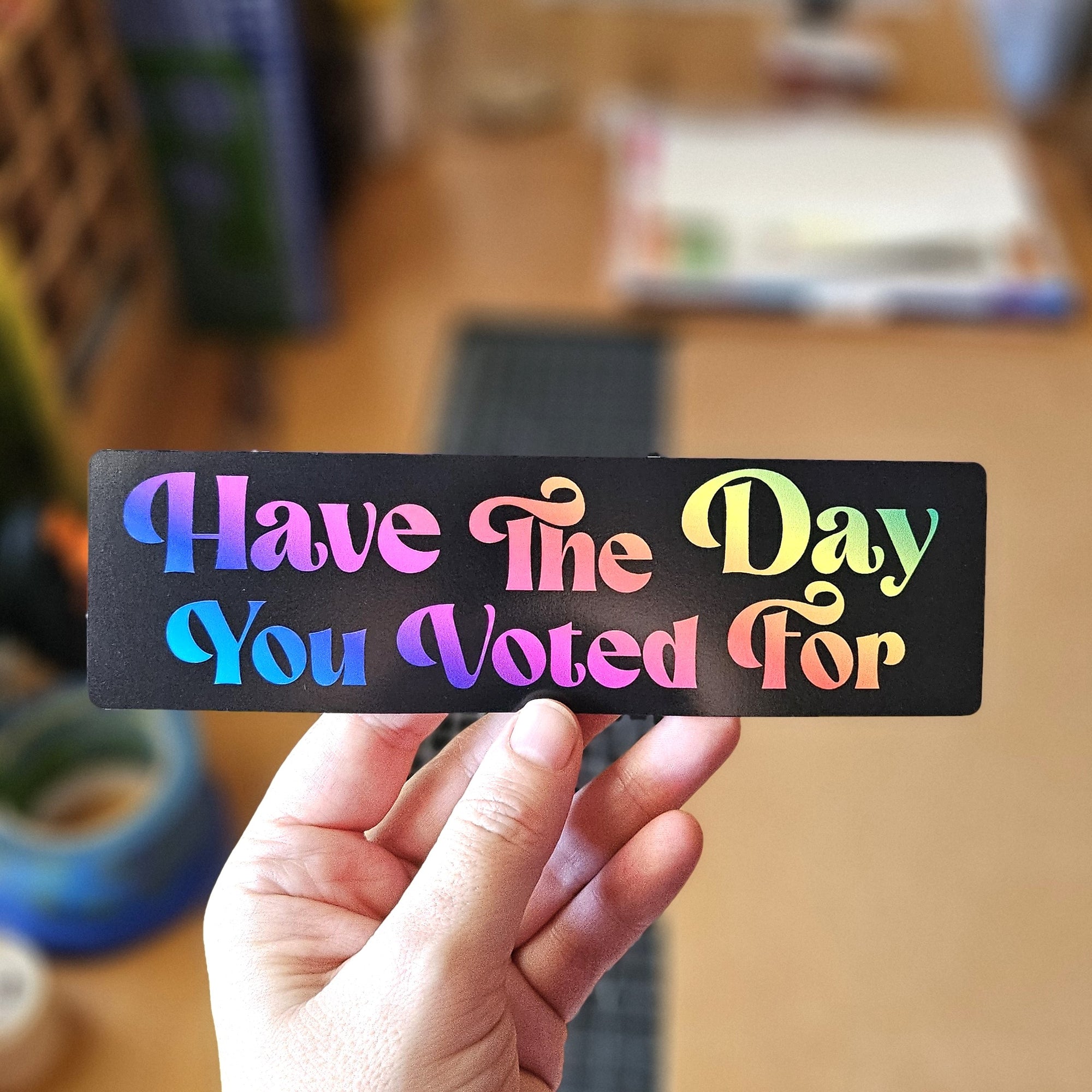 rectangular vinyl stickers that say “Have the Day You Voted For,” shown with  rainbow holographic-style text on black backgrounds.

