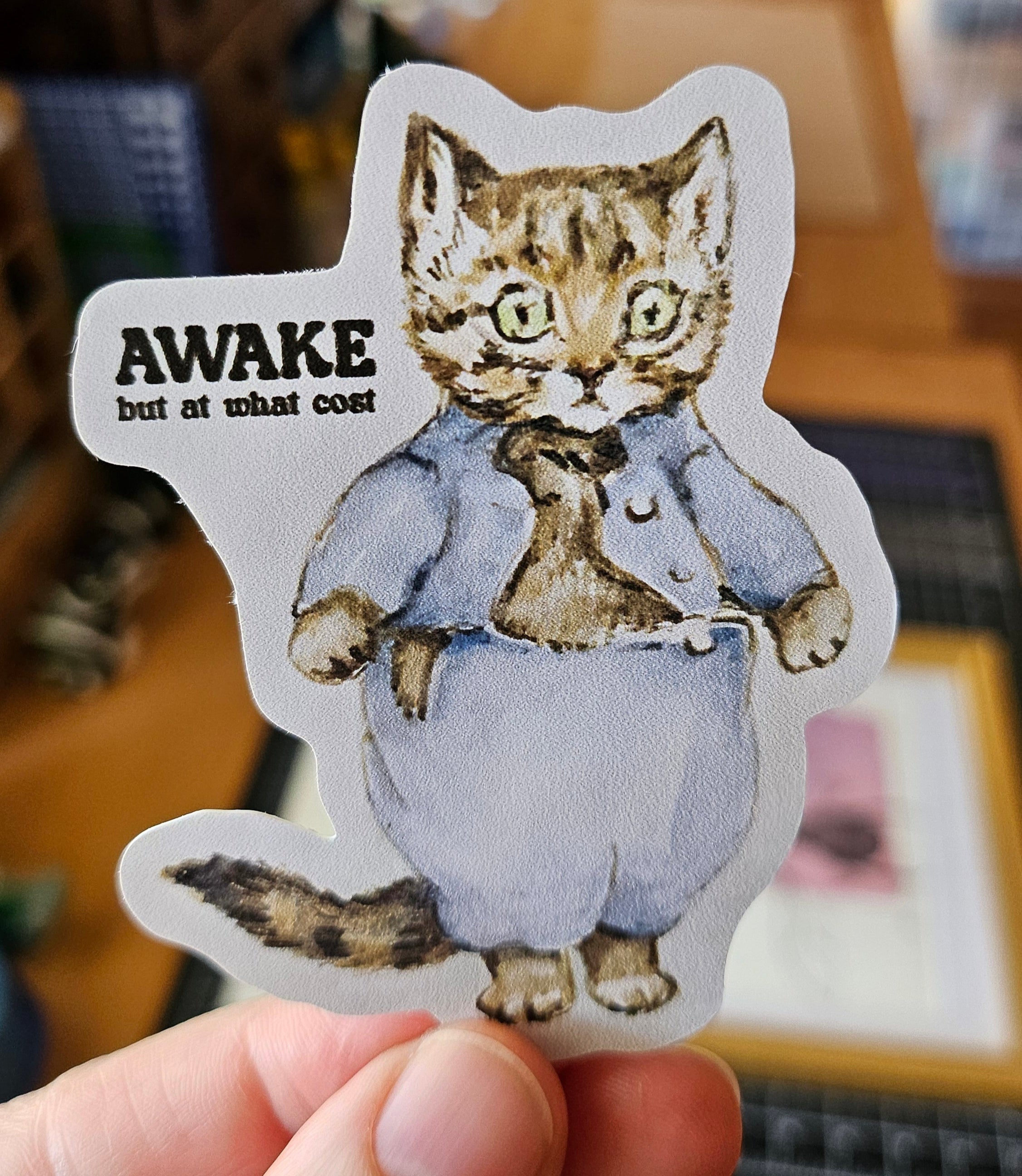 Die cut sticker featuring a vintage storybook-style cat wearing blue clothing with the phrase “Awake but at what cost.”
