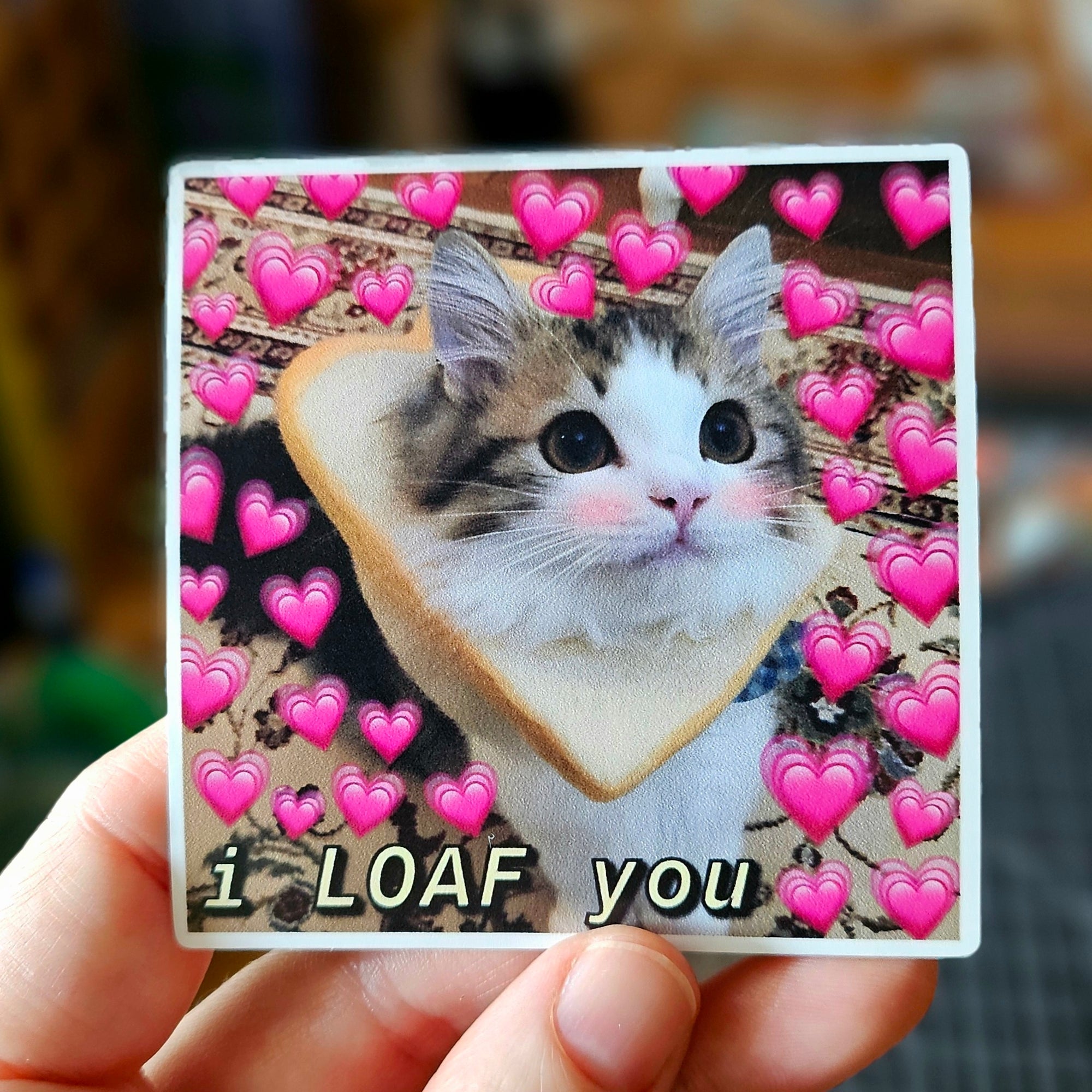 Square vinyl sticker featuring a cute cat wearing a slice of bread surrounded by pink hearts with the phrase “I Loaf You.”
