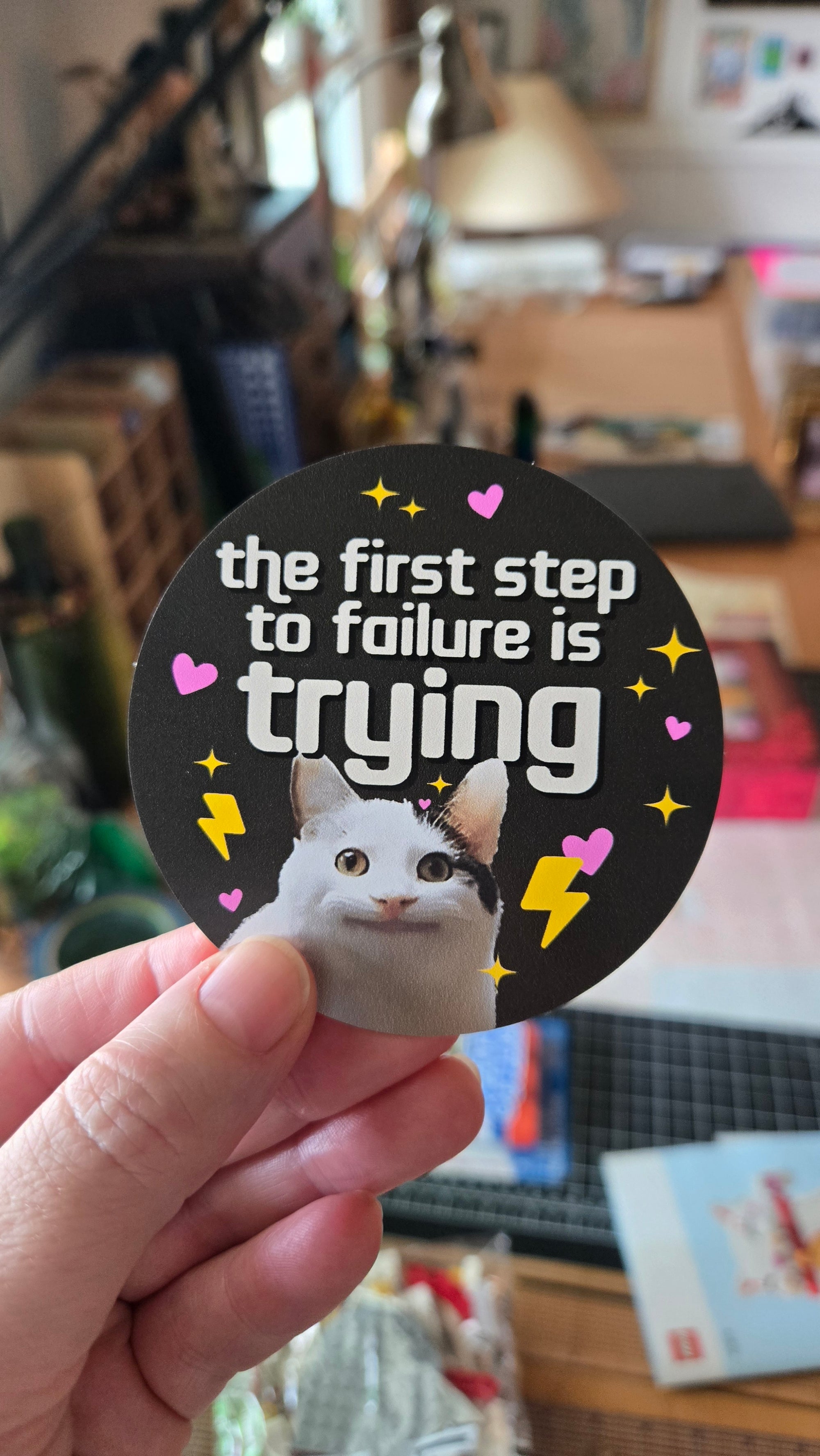 round vinyl sticker featuring a wide-eyed cat with hearts, sparkles, lightning bolts, and the phrase “the first step to failure is trying.”
