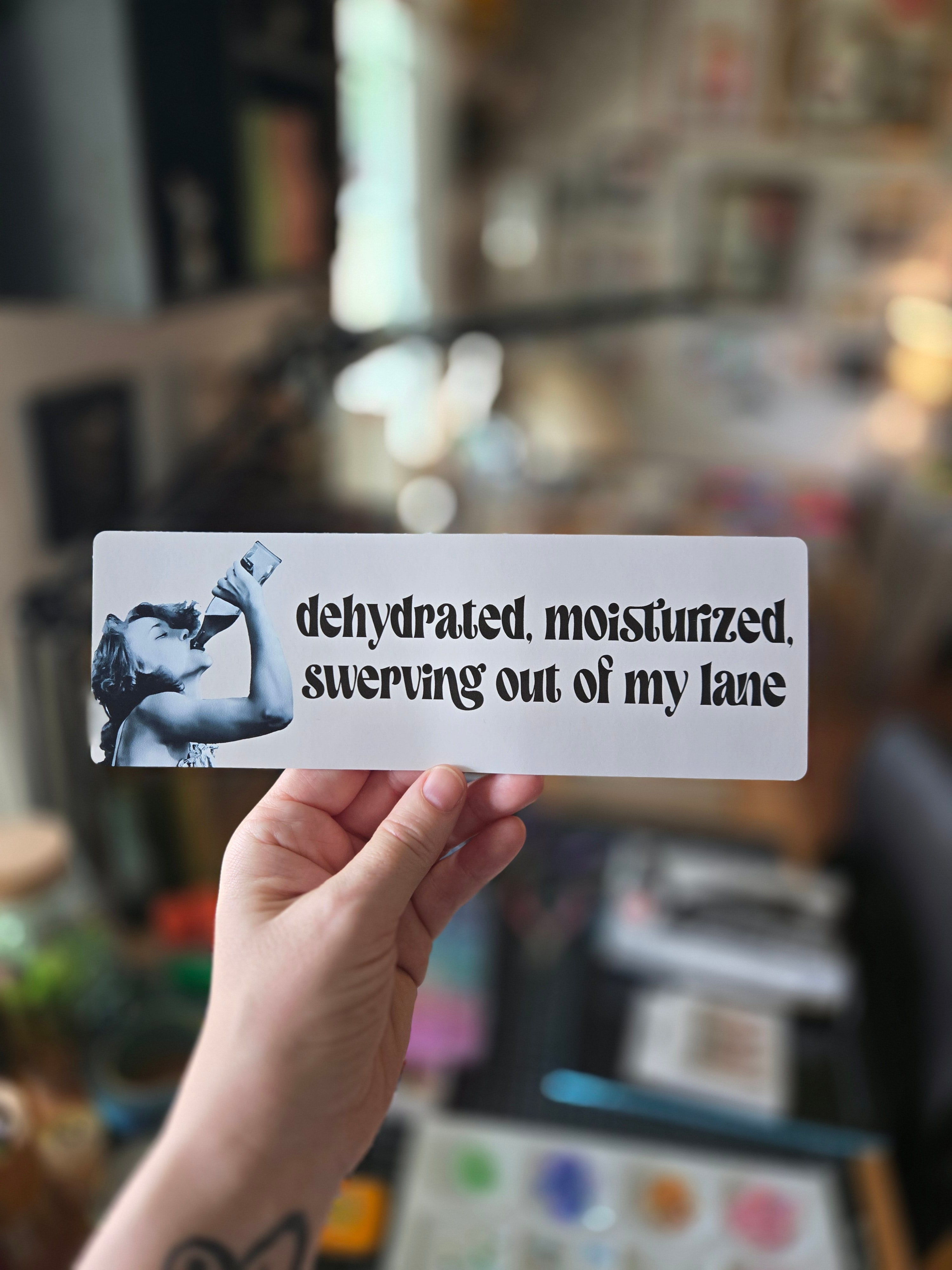 A vinyl sticker with the phrase 'dehydrated, moisturized, swerving out of my lane' printed on it, alongside an image of a person holding a drink.