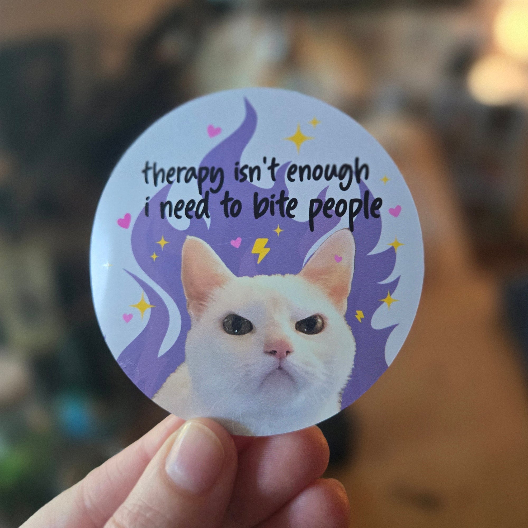 Round vinyl sticker featuring a serious white cat on a pastel background with hearts, sparkles, and the phrase “therapy isn’t enough i need to bite people.”
