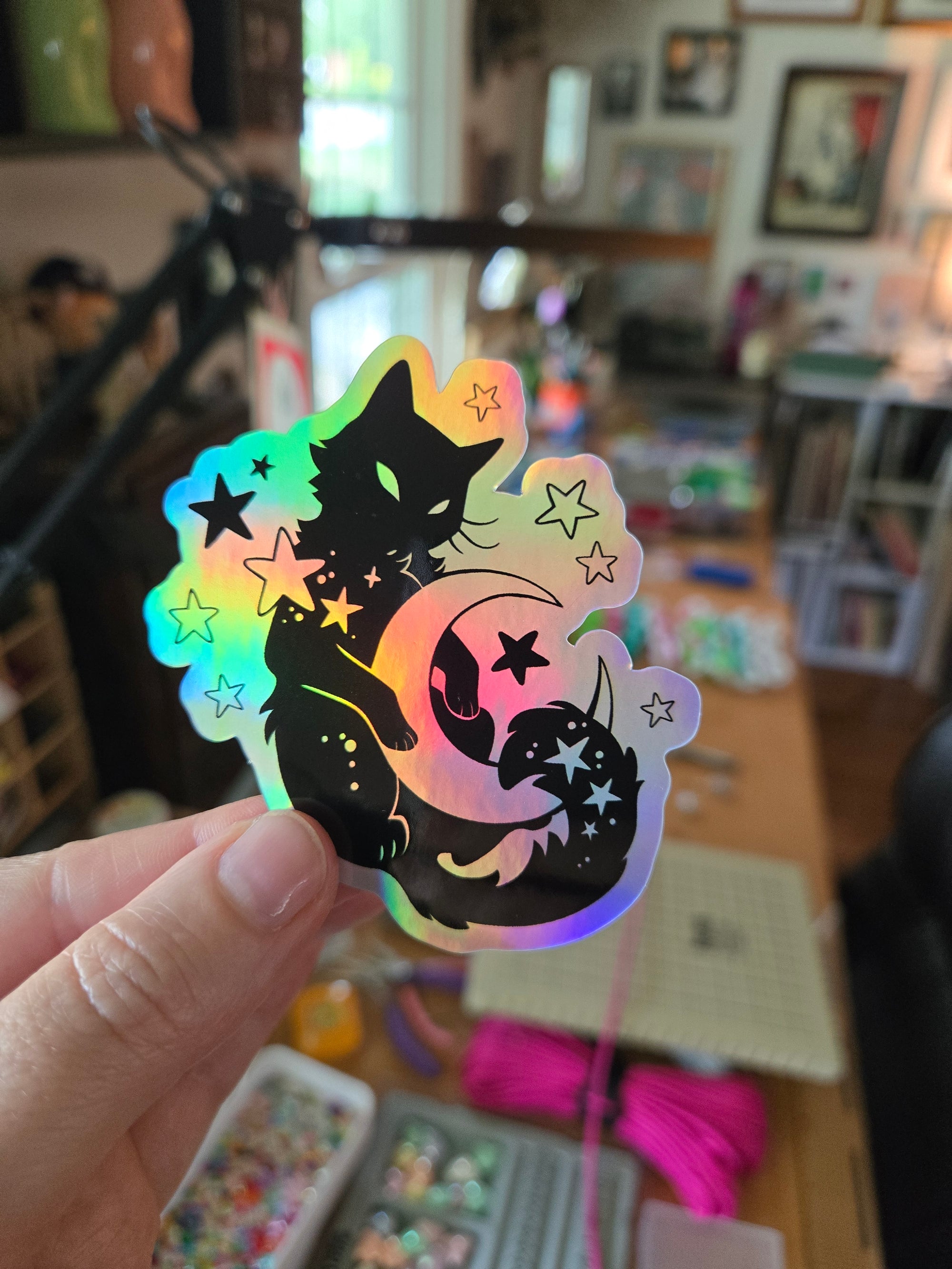 Die cut sticker featuring a black cat curled around a crescent moon with stars and a soft rainbow background.
