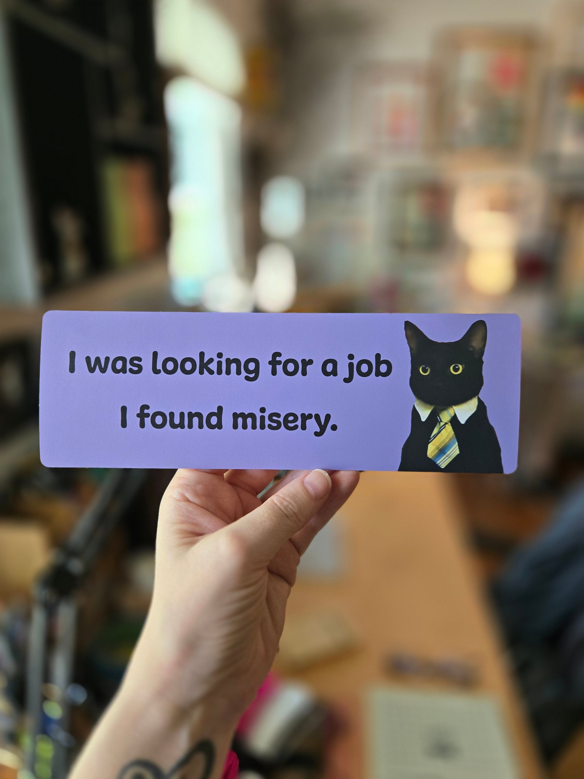 Purple bookmark with a cat illustration and humorous text held by a hand against a blurred background.