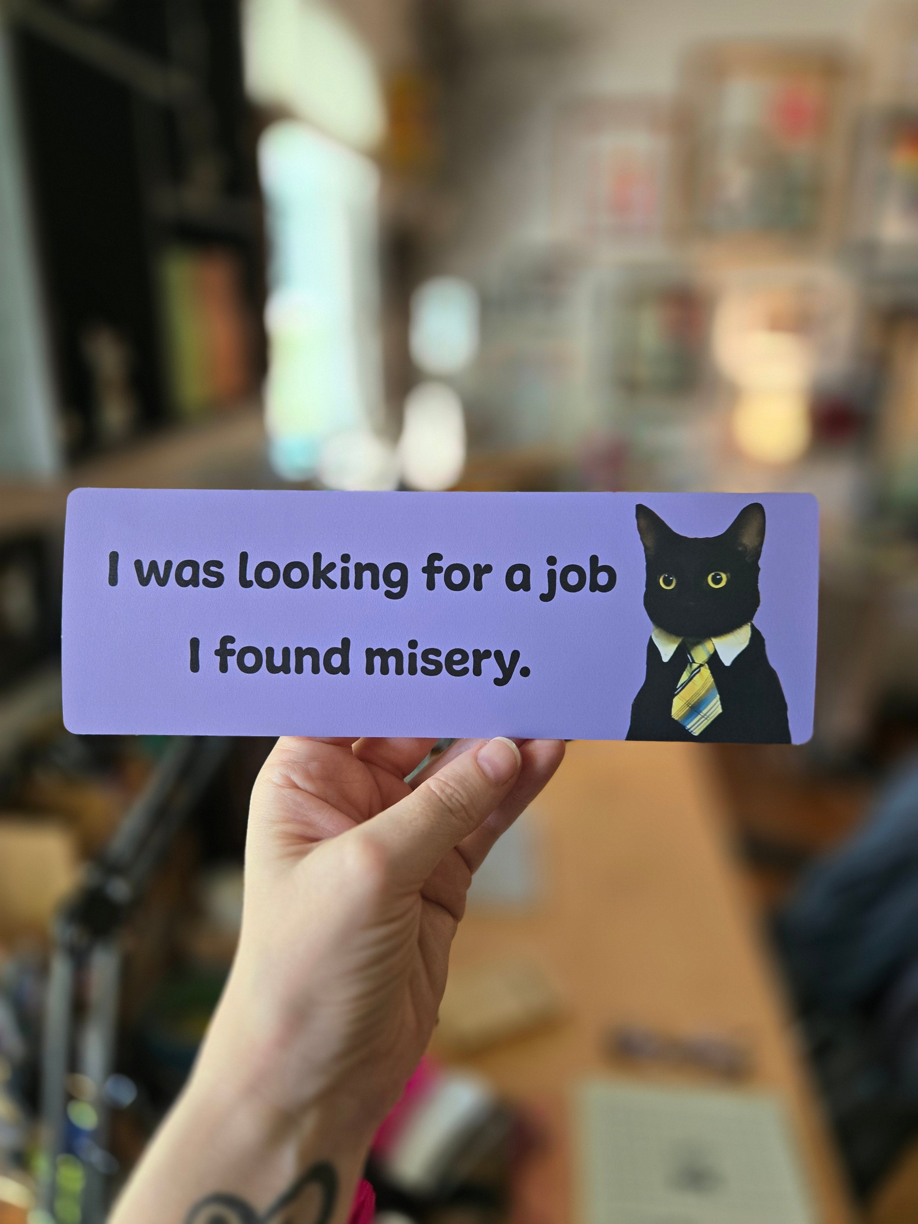 Purple bookmark with a cat illustration and humorous text held by a hand against a blurred background.