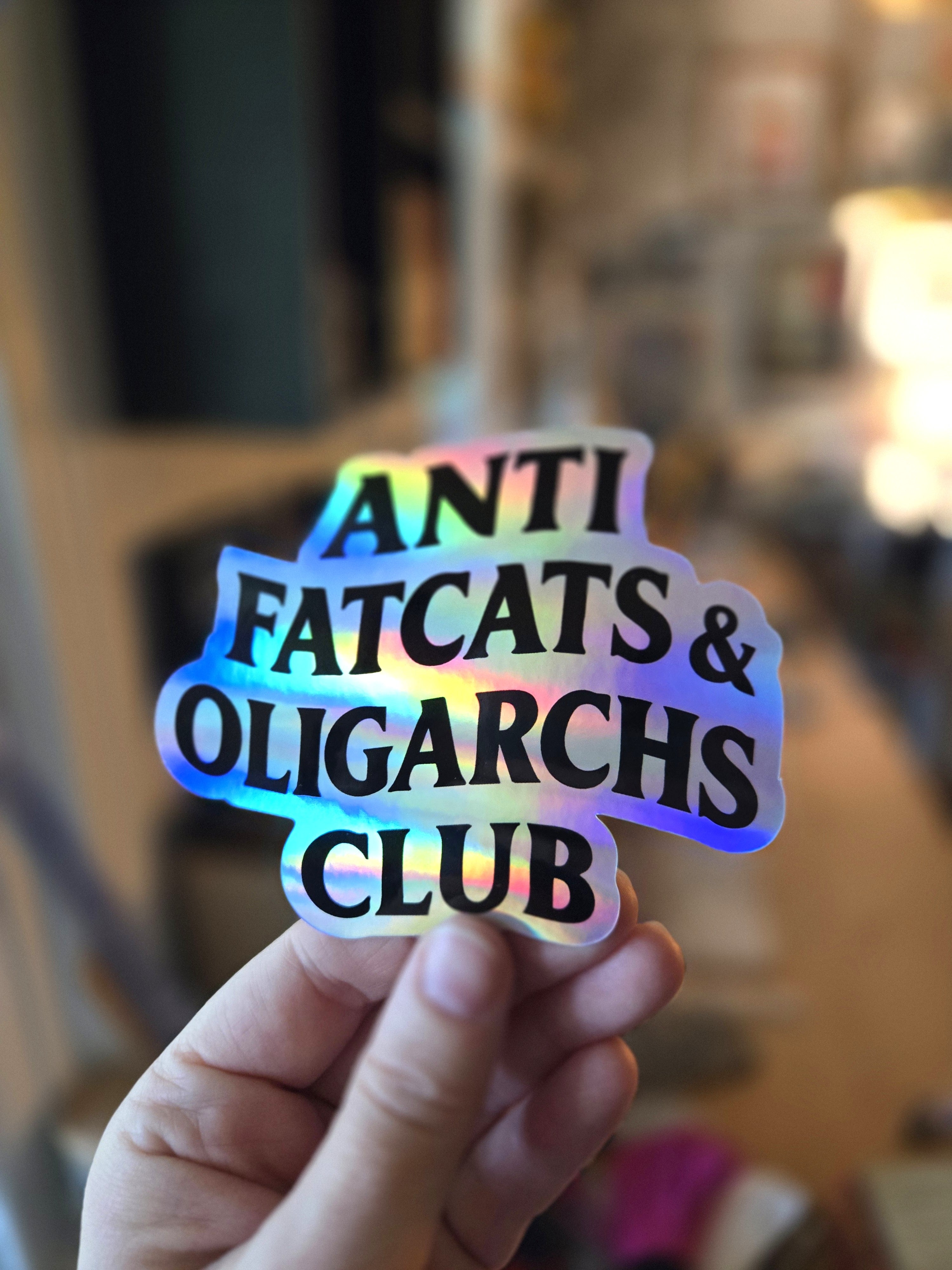 Holographic sticker with 'ANTI FATCATS & OLIGARCHS CLUB' text held by a hand against a blurred indoor background.