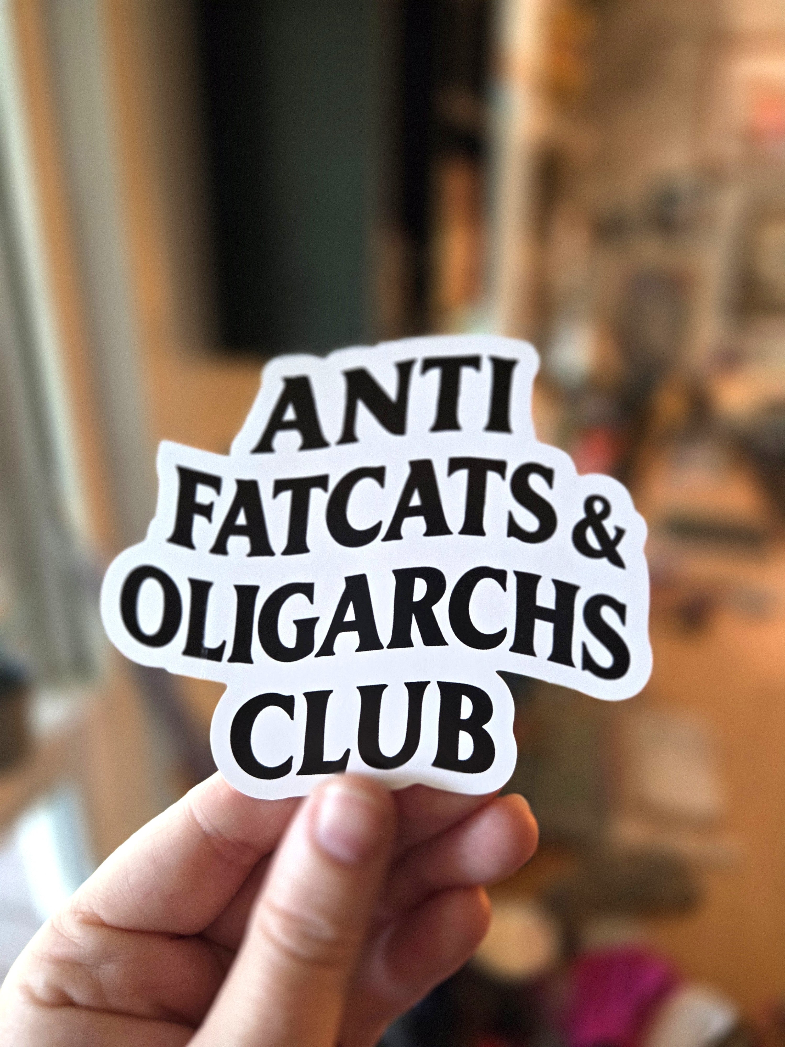 Sticker with 'ANTI FATCATS & OLIGARCHS CLUB' text held by a hand against a blurred indoor background.