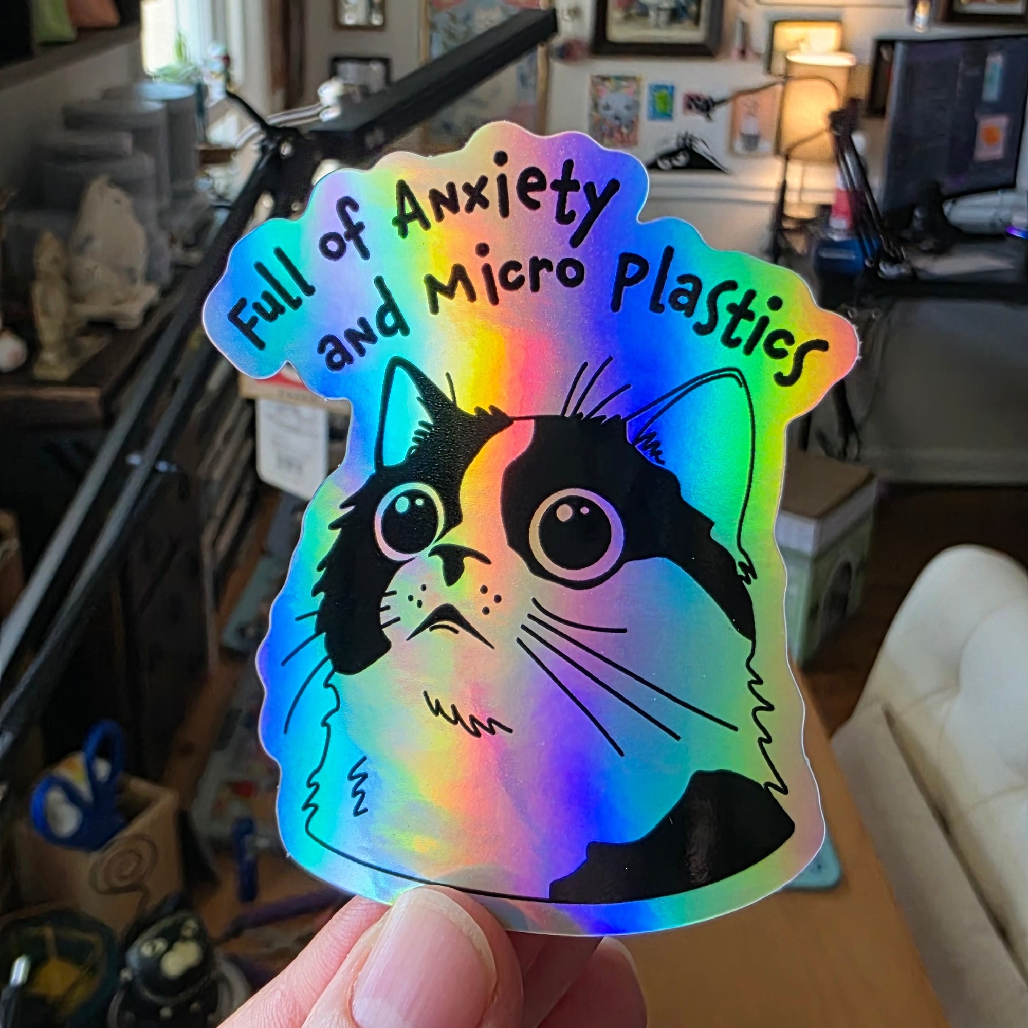 Holographic sticker with a cat design held by a hand in a cluttered room.