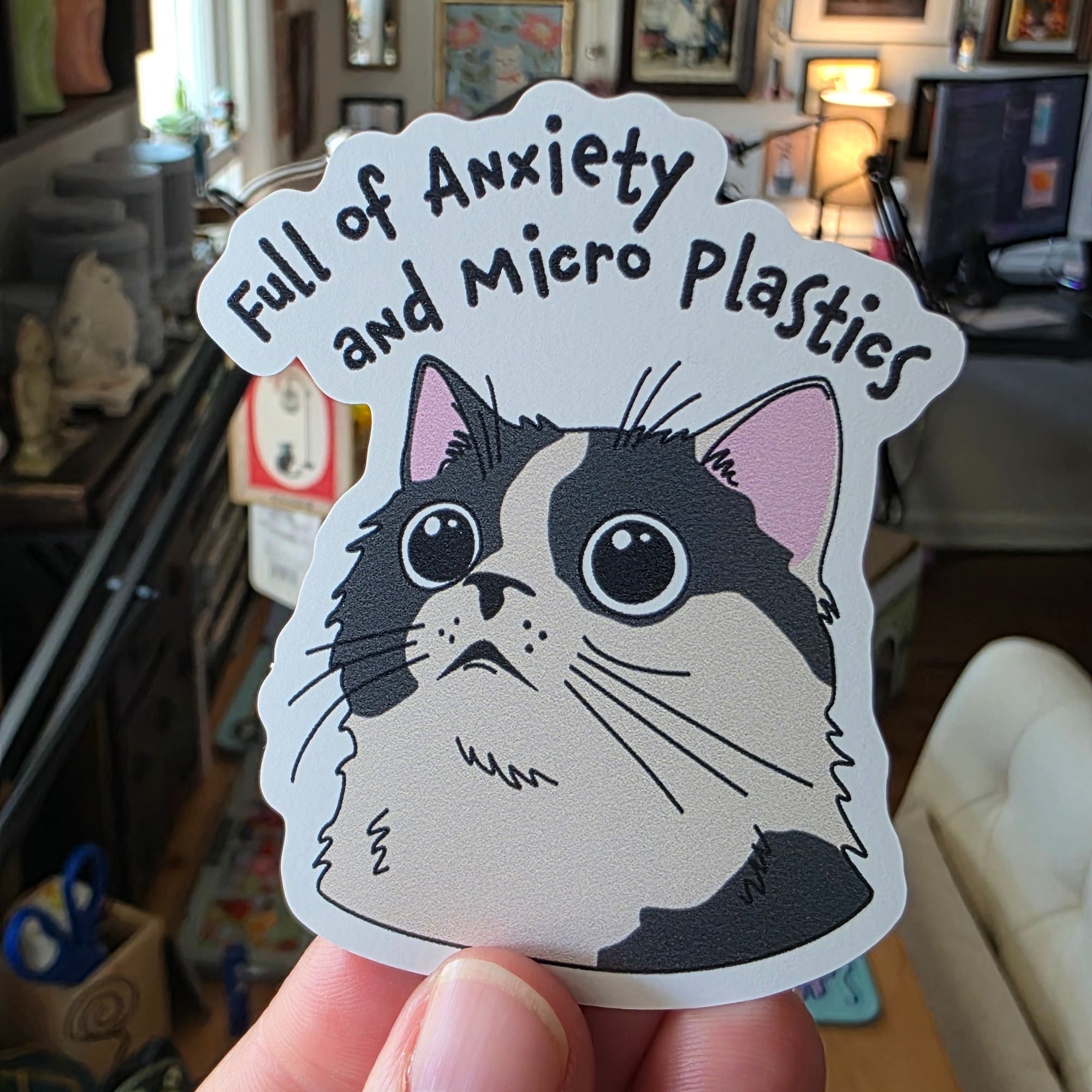 Hand holding a sticker of a cat with text in a cluttered room