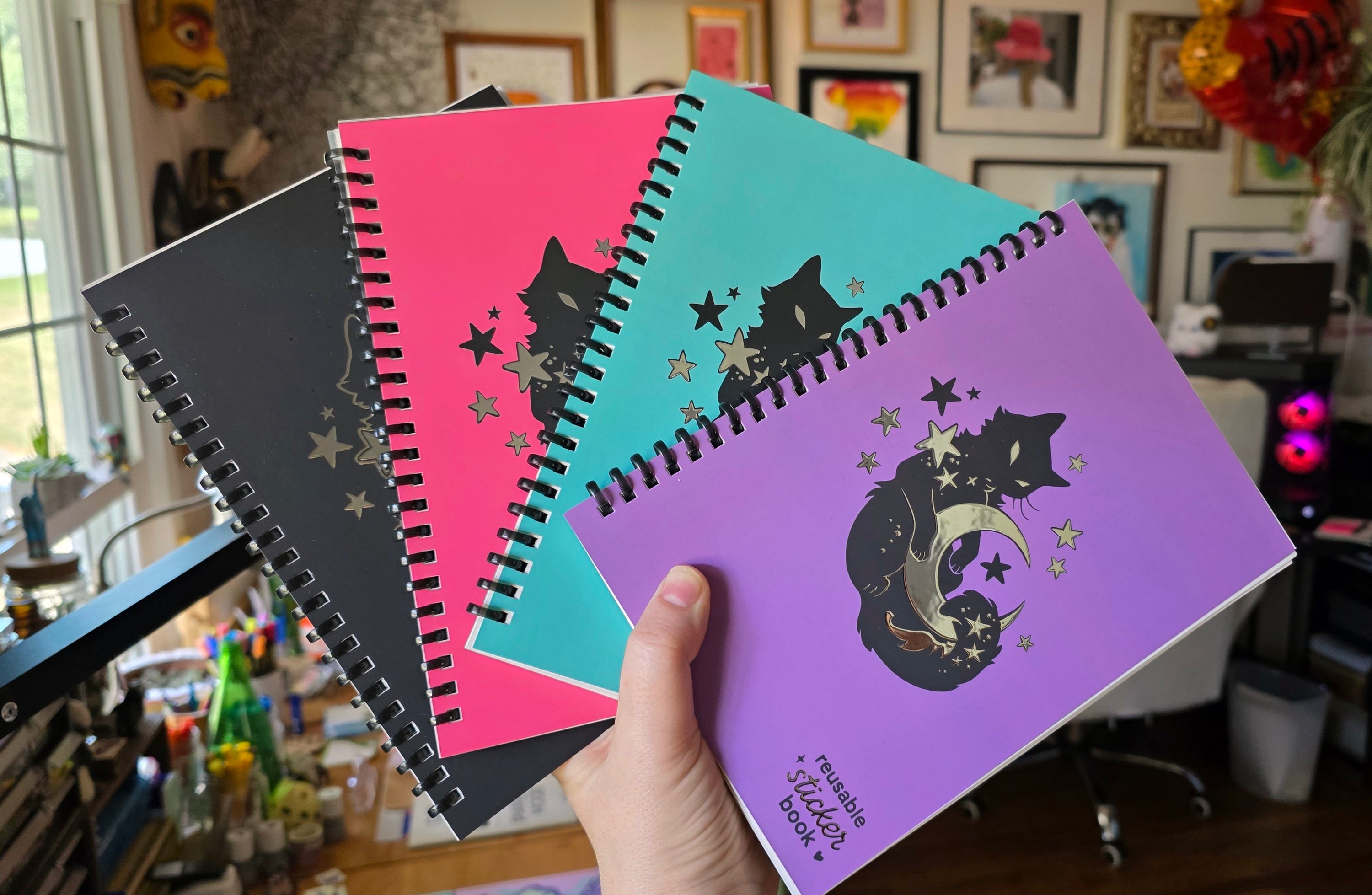 Hand holding four spiral-bound reusable sticker books in black, pink, teal, and purple, each featuring celestial cat artwork and silver foil details.
