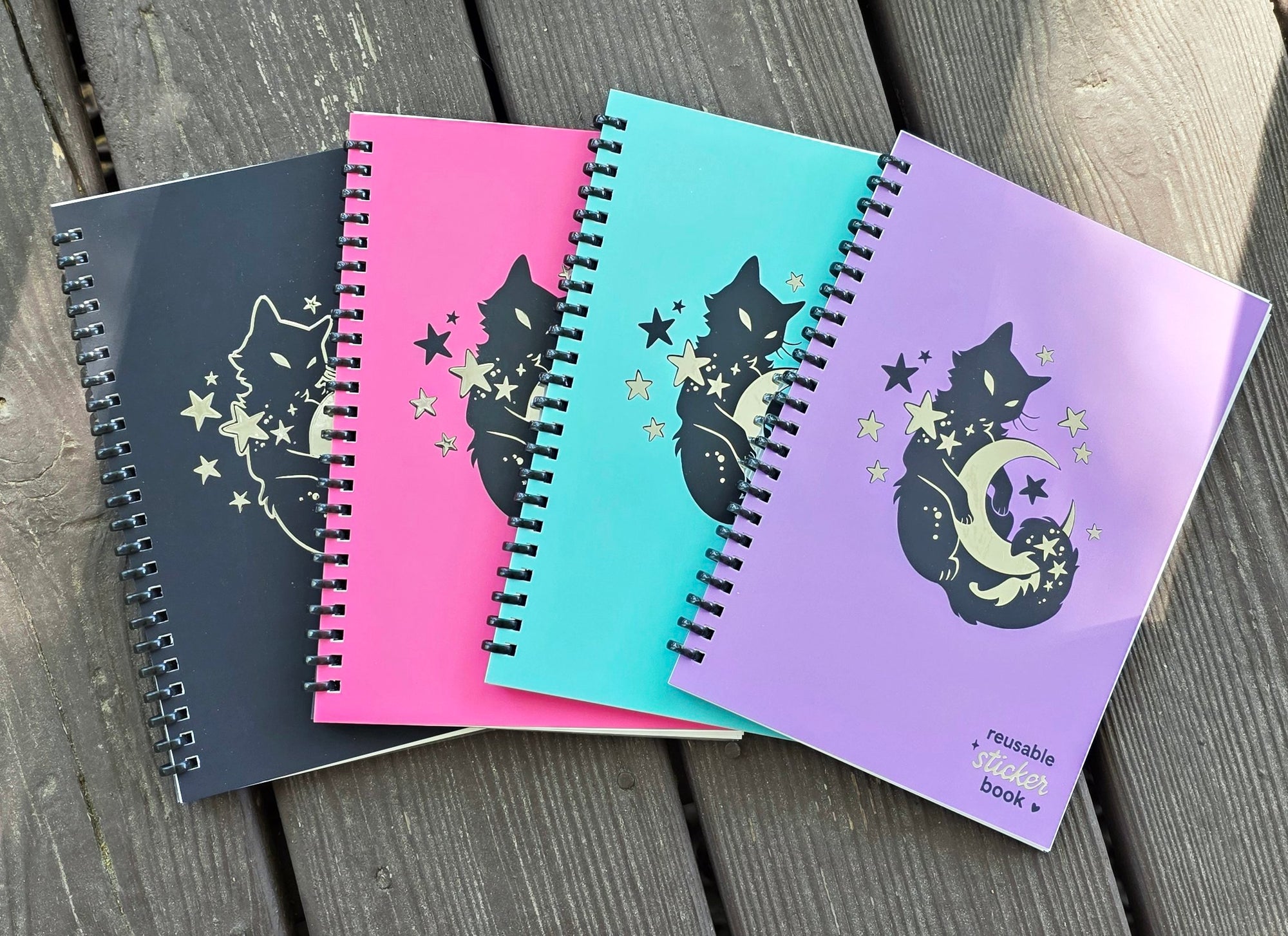 Four spiral-bound notebooks in different colors with cat illustrations on a wooden surface.